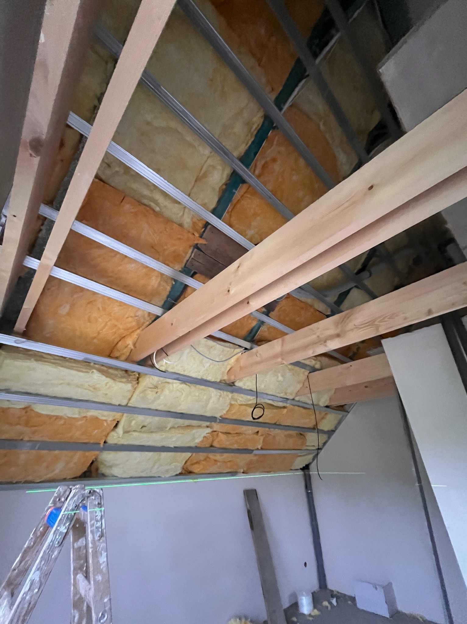 Skośne poddasze z izolacją z wełny mineralnej, widoczne drewniane belki i metalowe profile. Laserowy niwelator na drabinie wyznacza poziom. W tle fragment ściany.