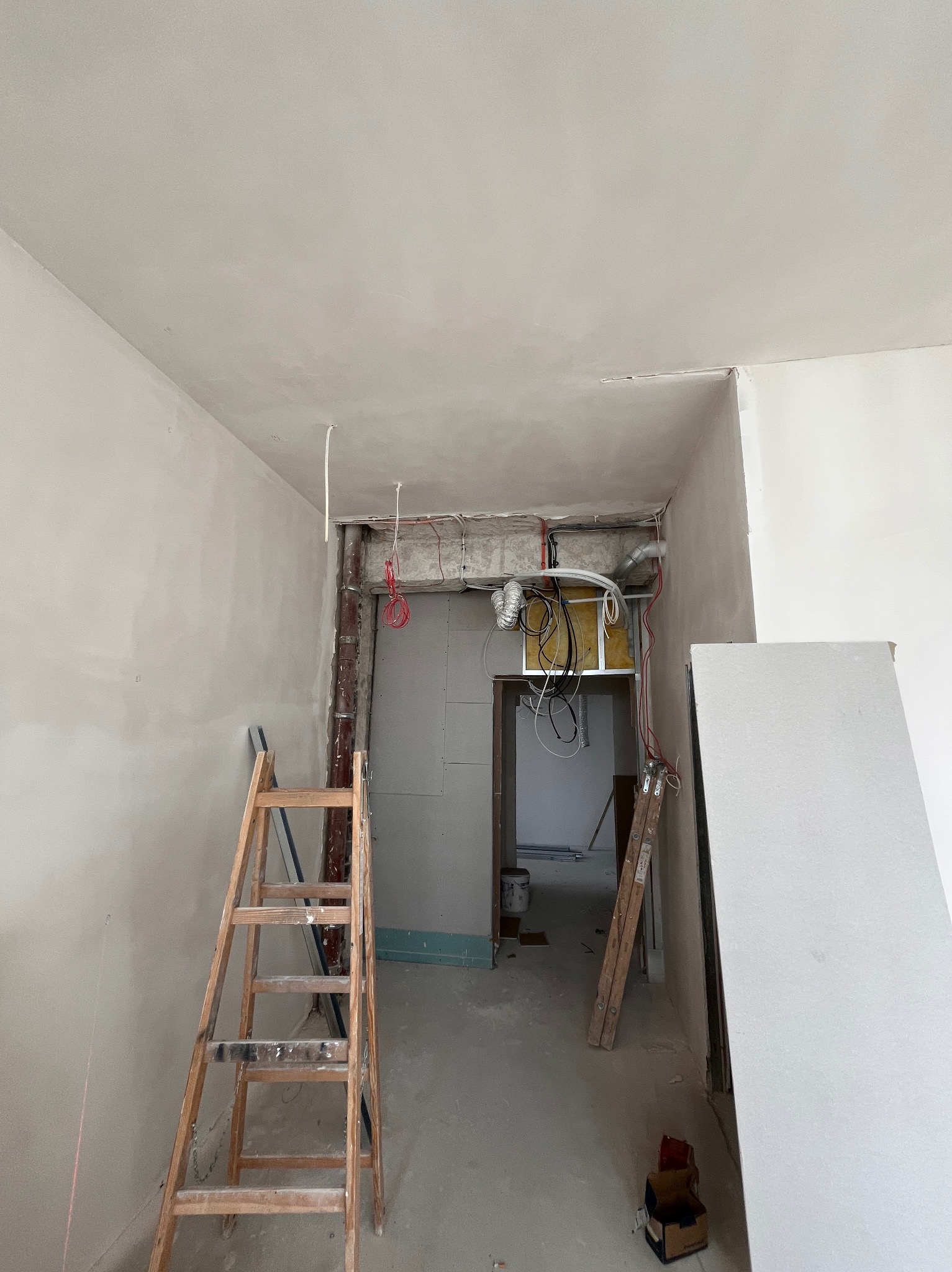 Wnętrze w trakcie prac wykończeniowych: ściany po szpachlowaniu, instalacje sufitowe, drabina oparta o ścianę, płyta gipsowa oparta o ścianę po prawej stronie.