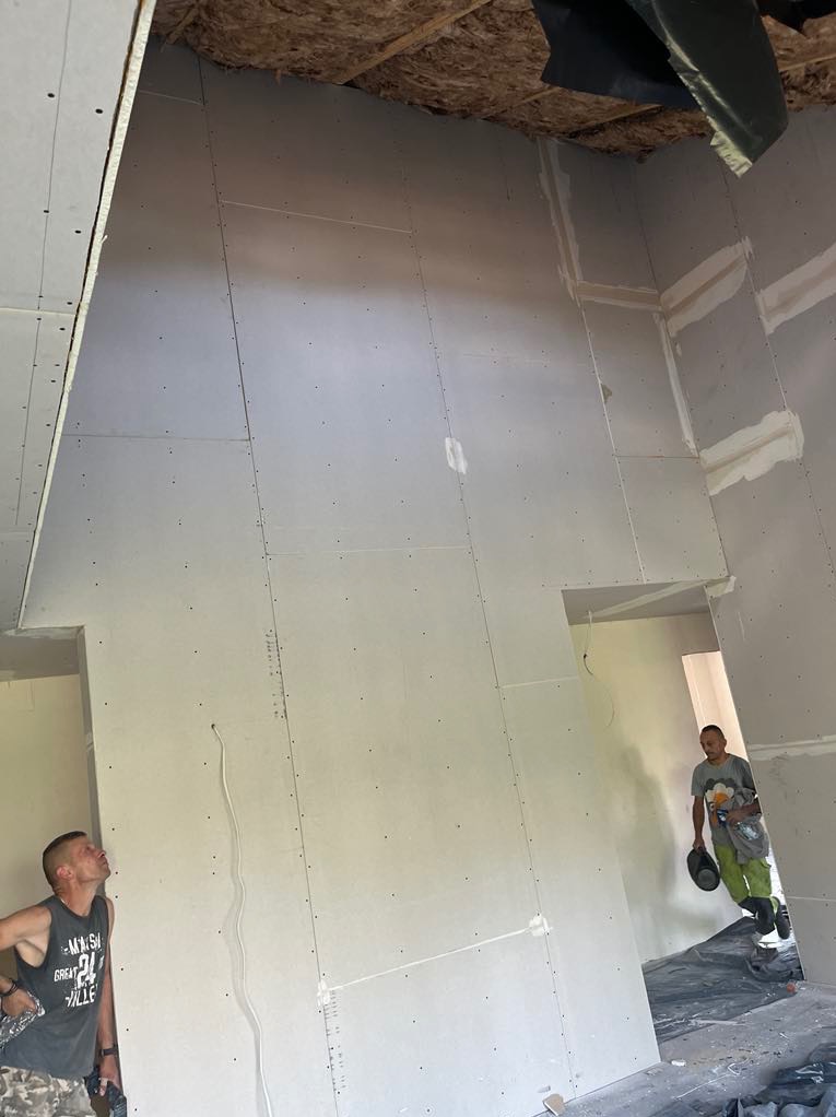 Montaż płyt gipsowo-kartonowych na ścianach i suficie wewnątrz budynku. Widoczne profile i izolacja na poddaszu. Dwóch mężczyzn w pomieszczeniu.