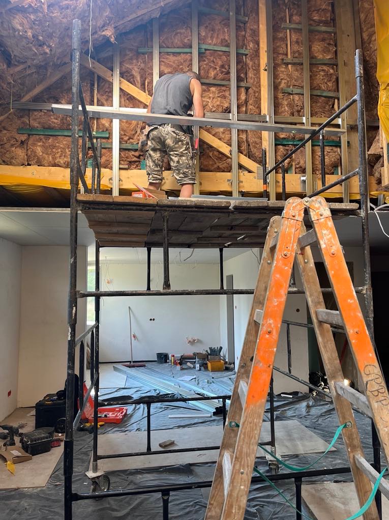 Pracownik na rusztowaniu montuje metalowy stelaż na ocieplonym poddaszu. W tle widoczne narzędzia i drabina, tworząc klimat remontowanego wnętrza.