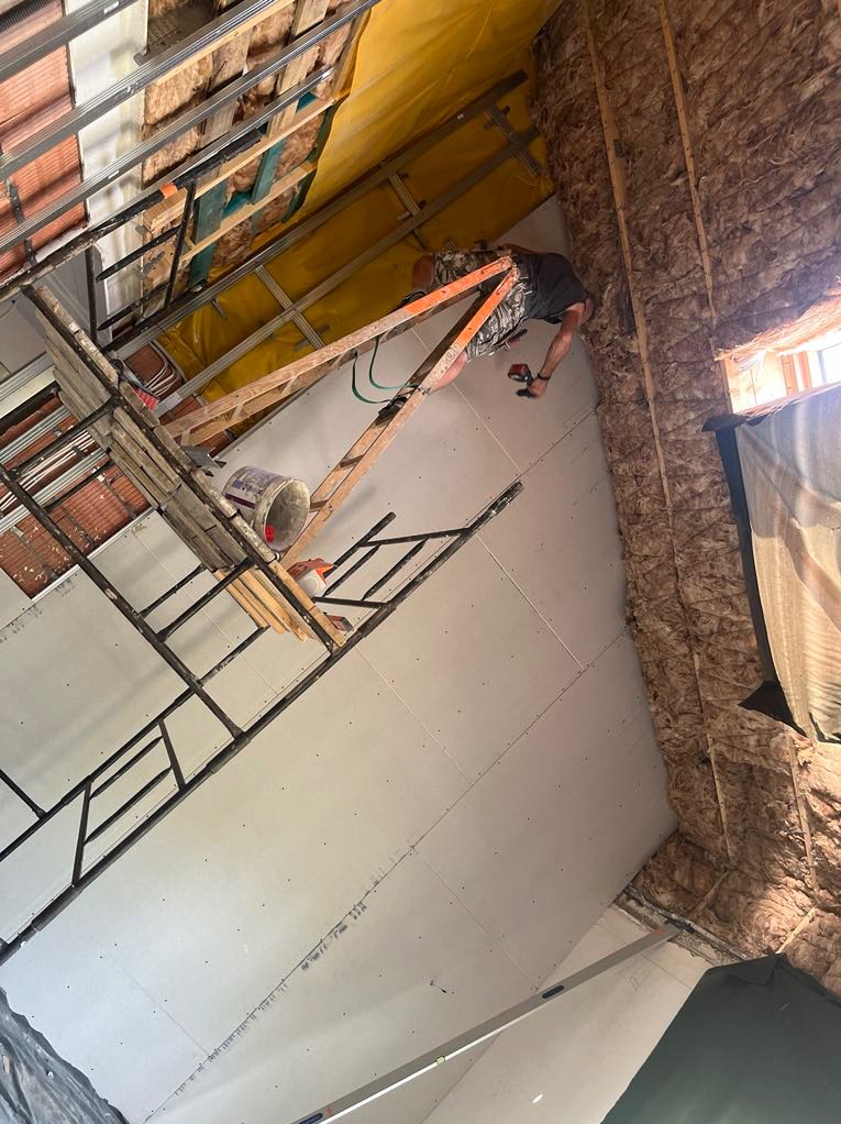 Montaż płyt na poddaszu: pracownik na drabinie mocuje płytę, widoczne ocieplenie, konstrukcja metalowa, narzędzia. Ujęcie z dołu.