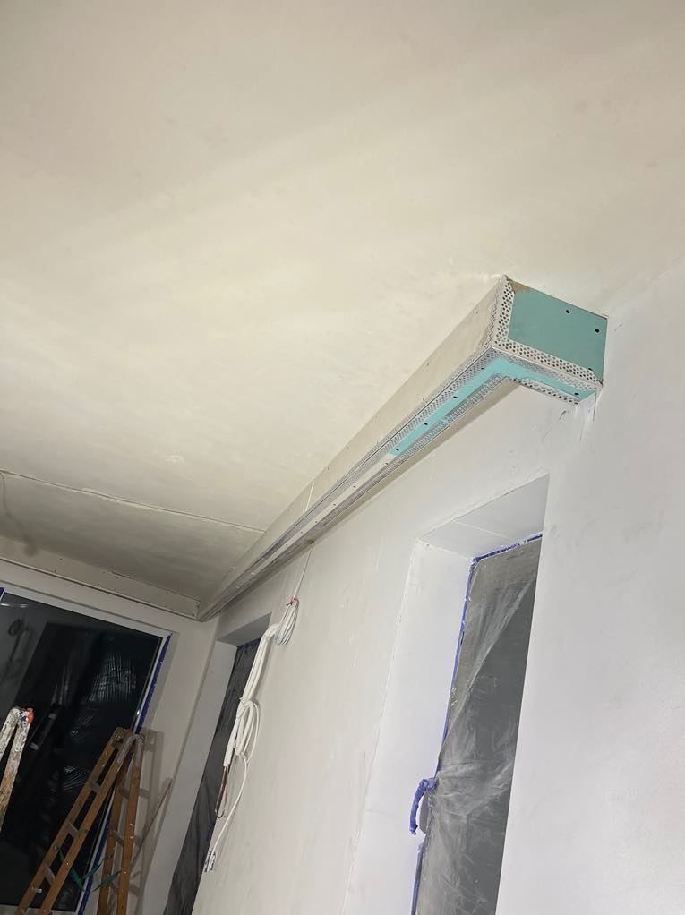 Narożnik sufitowy z płyty KG, widoczne profile i siatka wzmacniająca. Ściany świeżo pomalowane na biało, zabezpieczone okno folią malarską. Prace wykończeniowe w trakcie.