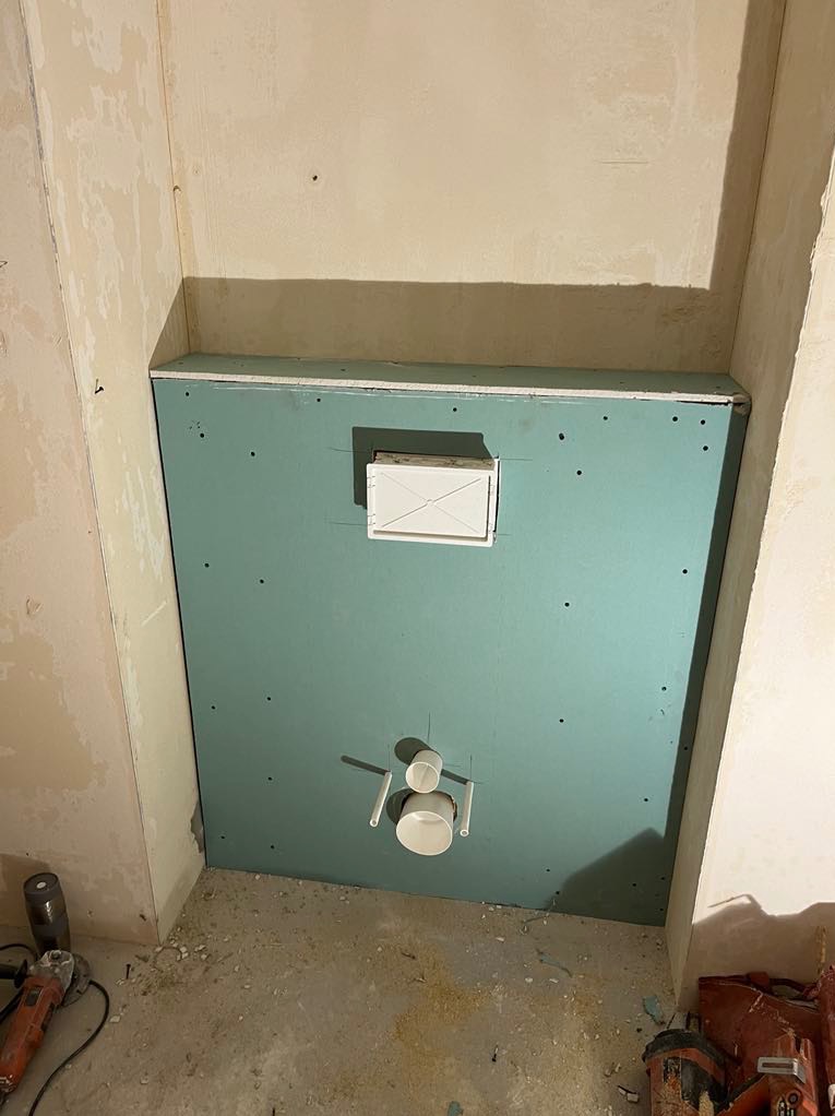 Zielona płyta GK z wyciętymi otworami na instalacje, zamontowana w narożniku pomieszczenia w trakcie prac wykończeniowych. Widoczne narzędzia.