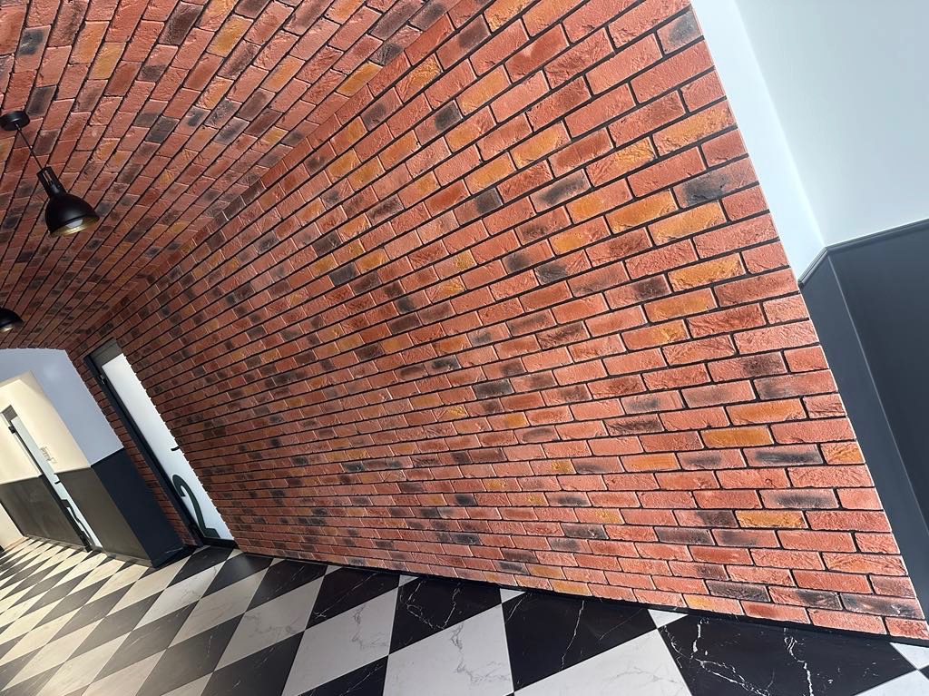 Ściana z cegieł dekoracyjnych w odcieniach czerwieni i brązu, ułożona pod kątem, z czarno-białą podłogą w szachownicę i widocznym oświetleniem sufitowym.