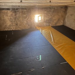 Cellu - Marcin Korzyński - Poddasze z surową cegłą, częściowo wyłożone czarnymi płytami izolacyjnymi i żółtą folią, oświetlone przez małe okno. Widoczne przewody pod sufitem.