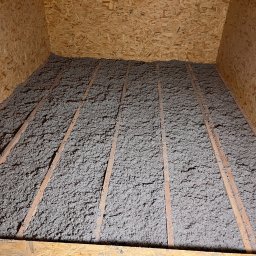 Cellu - Marcin Korzyński - Szara izolacja z wełny mineralnej wypełnia przestrzeń poddasza między drewnianymi belkami, widoczne płyty OSB na ścianach i suficie.