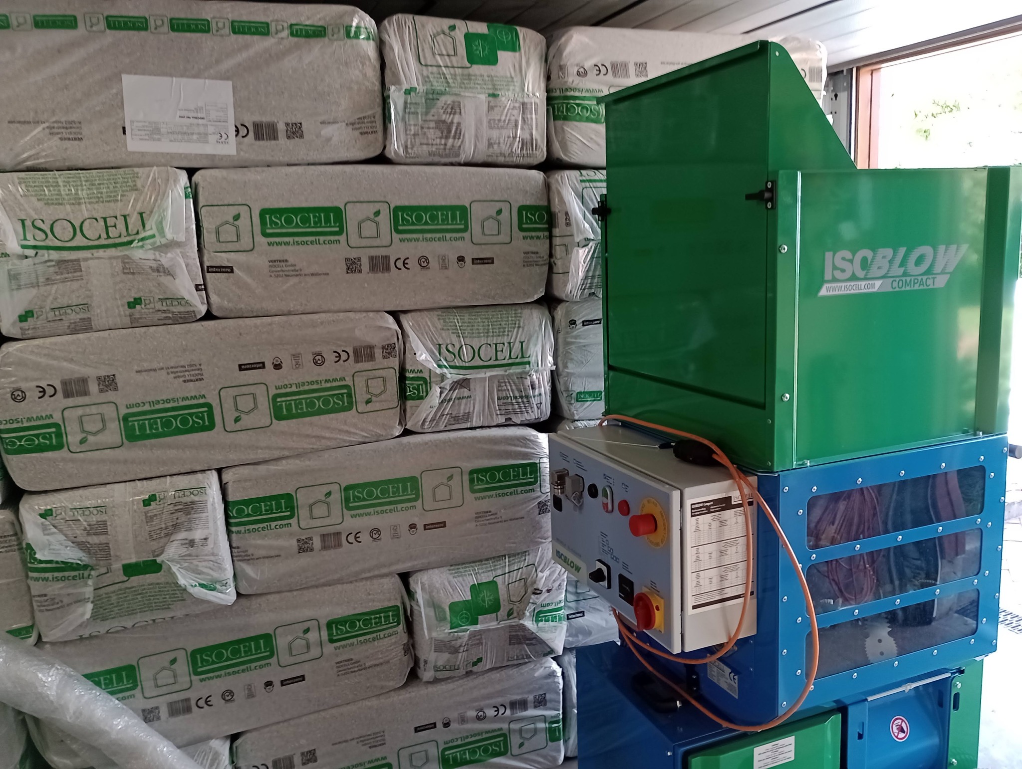 Stos worków z wełną celulozową Isocell obok maszyny do wdmuchiwania izolacji Isoblow Compact, przygotowanych do ocieplenia poddasza lub ścian.