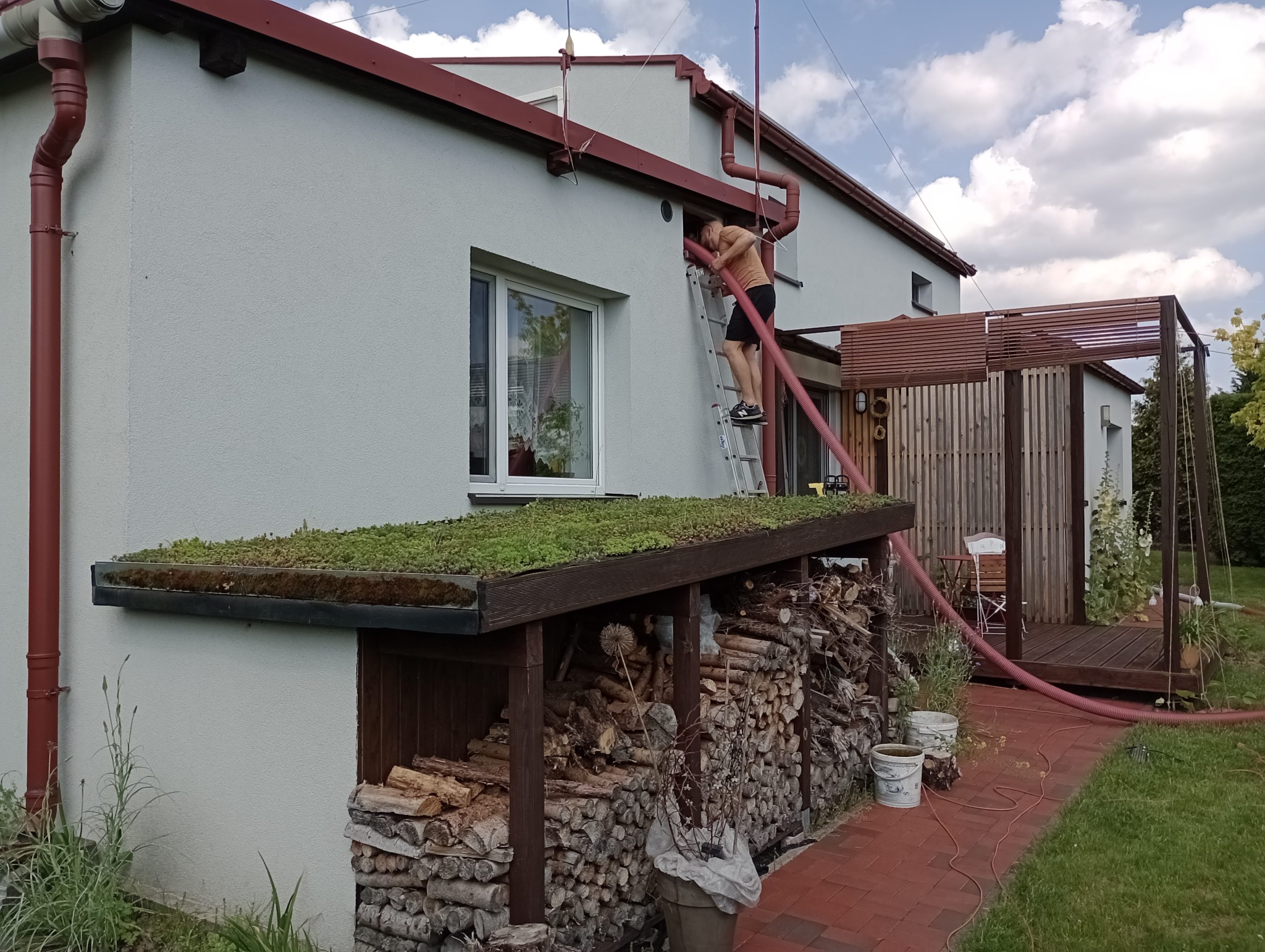 Pracownik na drabinie wprowadza rurę do otworu w ścianie domu, prawdopodobnie w celu wdmuchiwania materiału izolacyjnego; widoczny skład drewna z zielonym dachem.