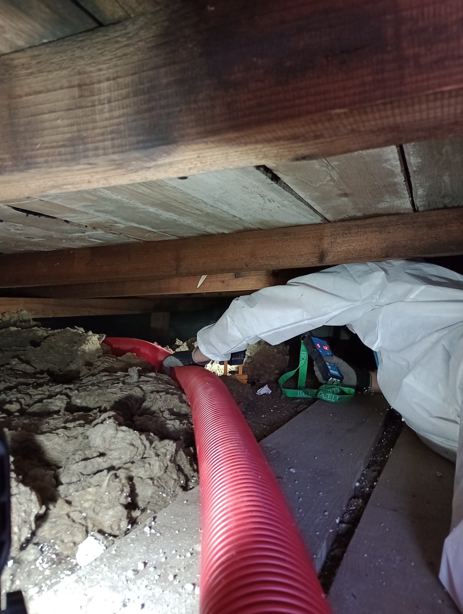 Pracownik w kombinezonie ochronnym w Tarnowskich Górach aplikuje izolację do poddasza za pomocą czerwonej rury, widoczne drewniane elementy konstrukcji dachu.