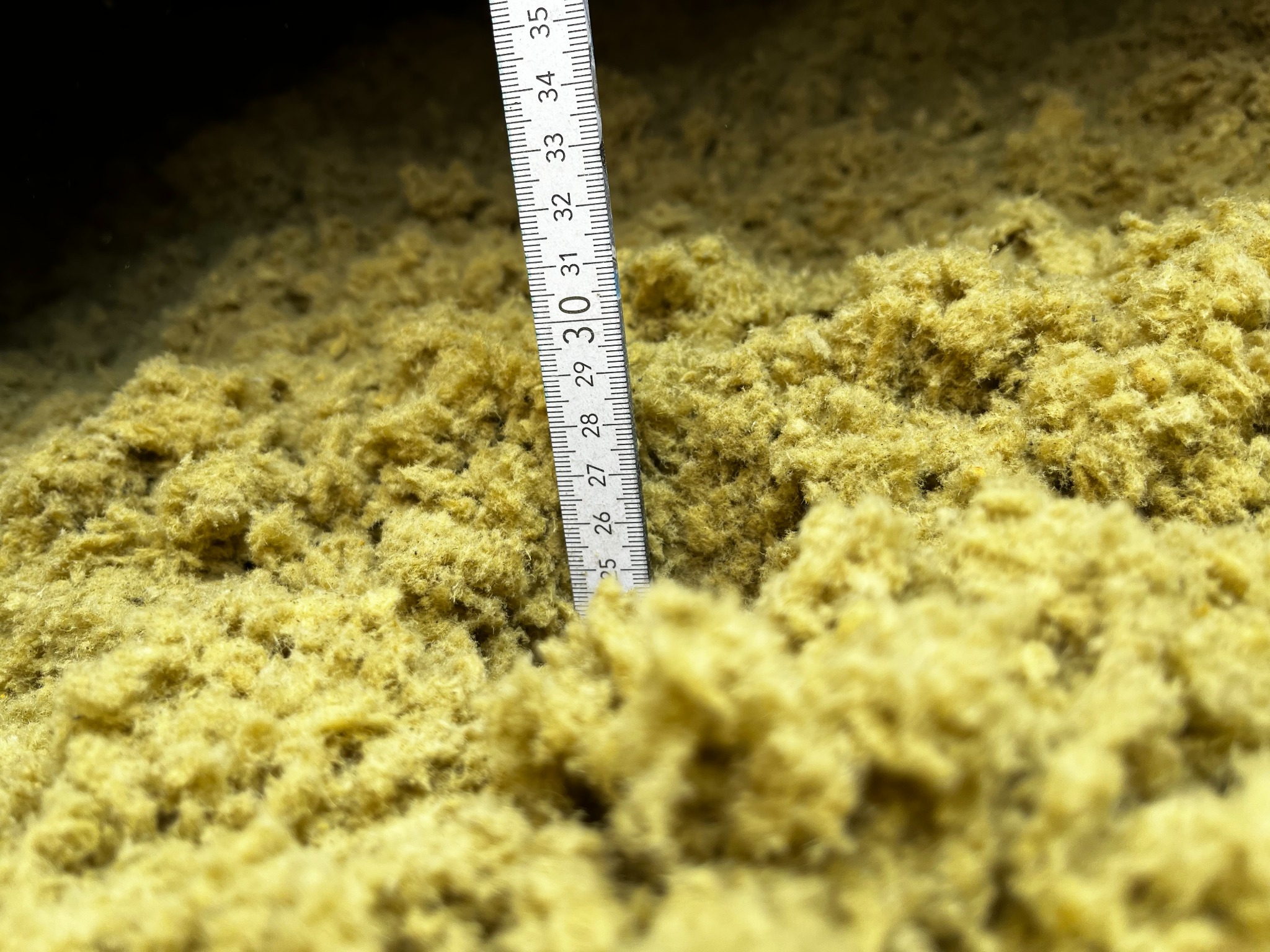 Pomiar grubości warstwy izolacyjnej, prawdopodobnie wełny, za pomocą rozkładanej miarki. Widoczny zakres od 25 do 35 cm.