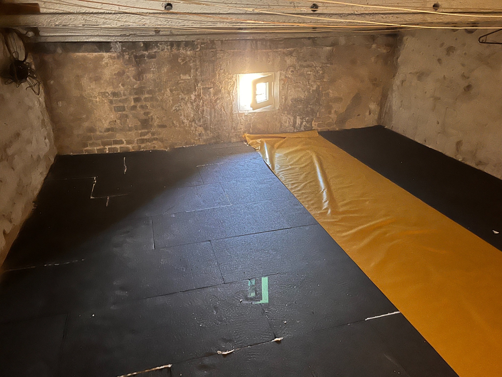 Poddasze z surową cegłą, częściowo wyłożone czarnymi płytami izolacyjnymi i żółtą folią, oświetlone przez małe okno. Widoczne przewody pod sufitem.