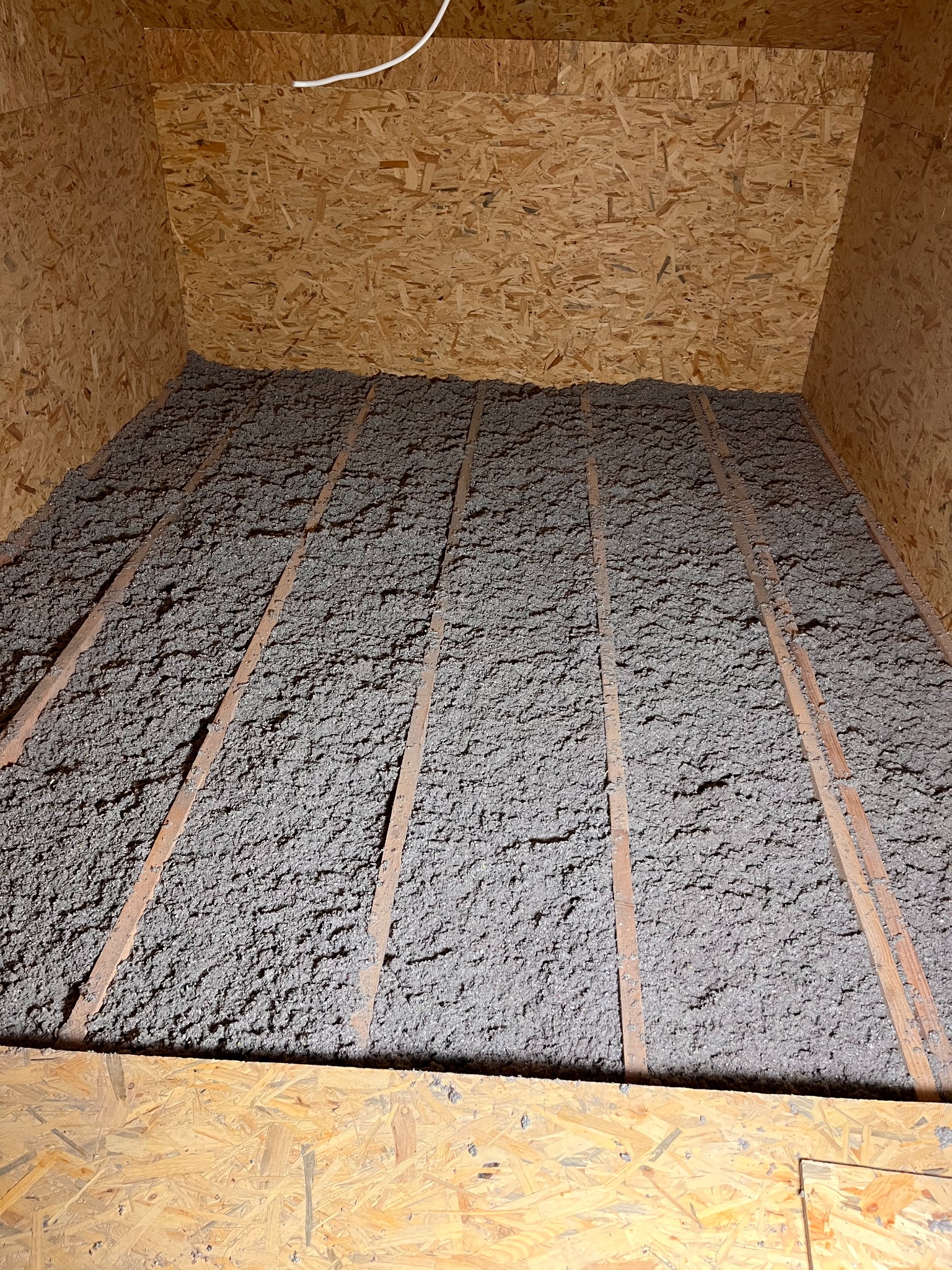 Szara izolacja z wełny mineralnej wypełnia przestrzeń poddasza między drewnianymi belkami, widoczne płyty OSB na ścianach i suficie.