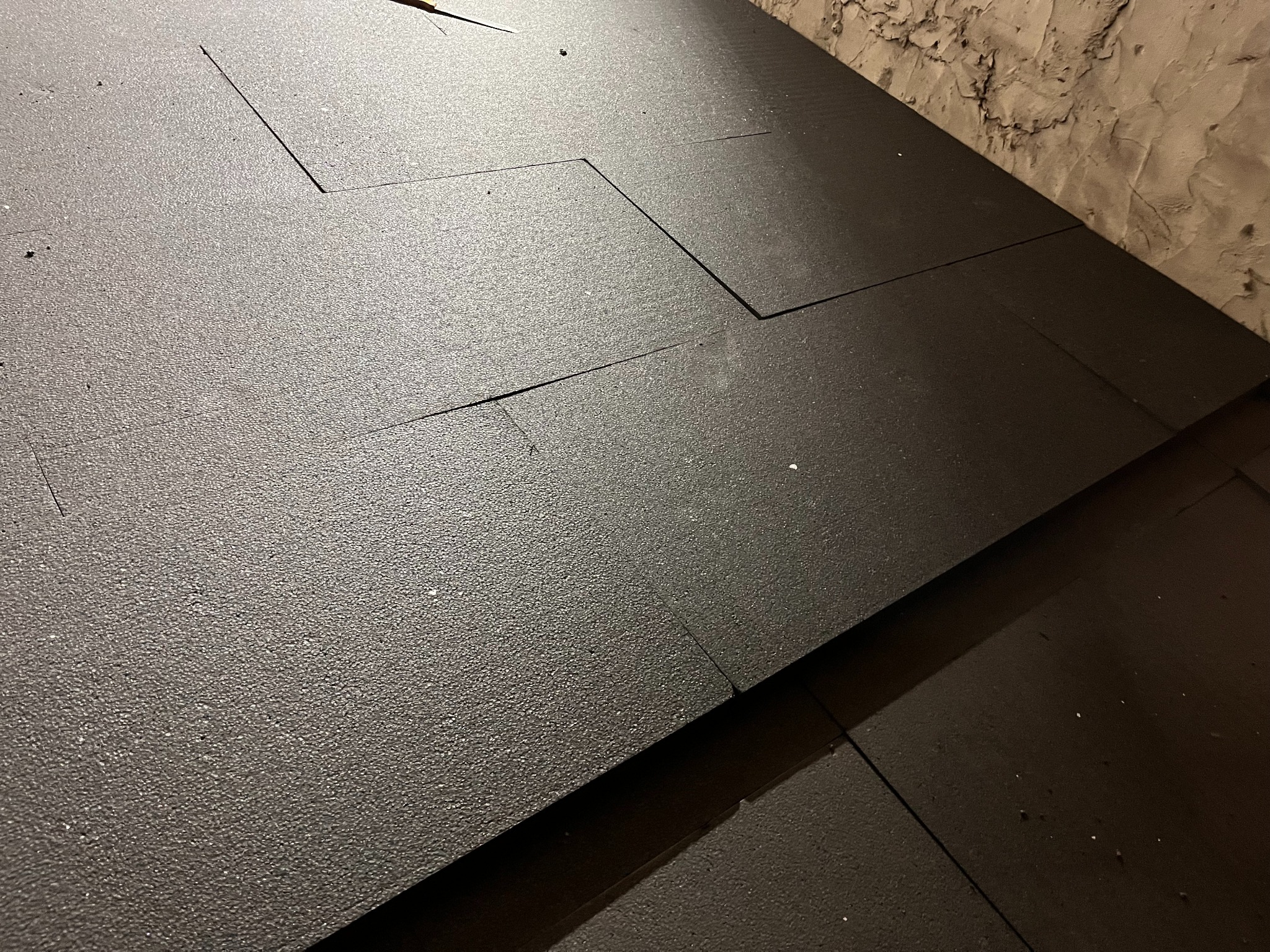 Ciemnoszare płyty izolacyjne ułożone na poddaszu, widoczne szczeliny między płytami i fragment ściany z nierówną fakturą. Izolacja termiczna w trakcie montażu.