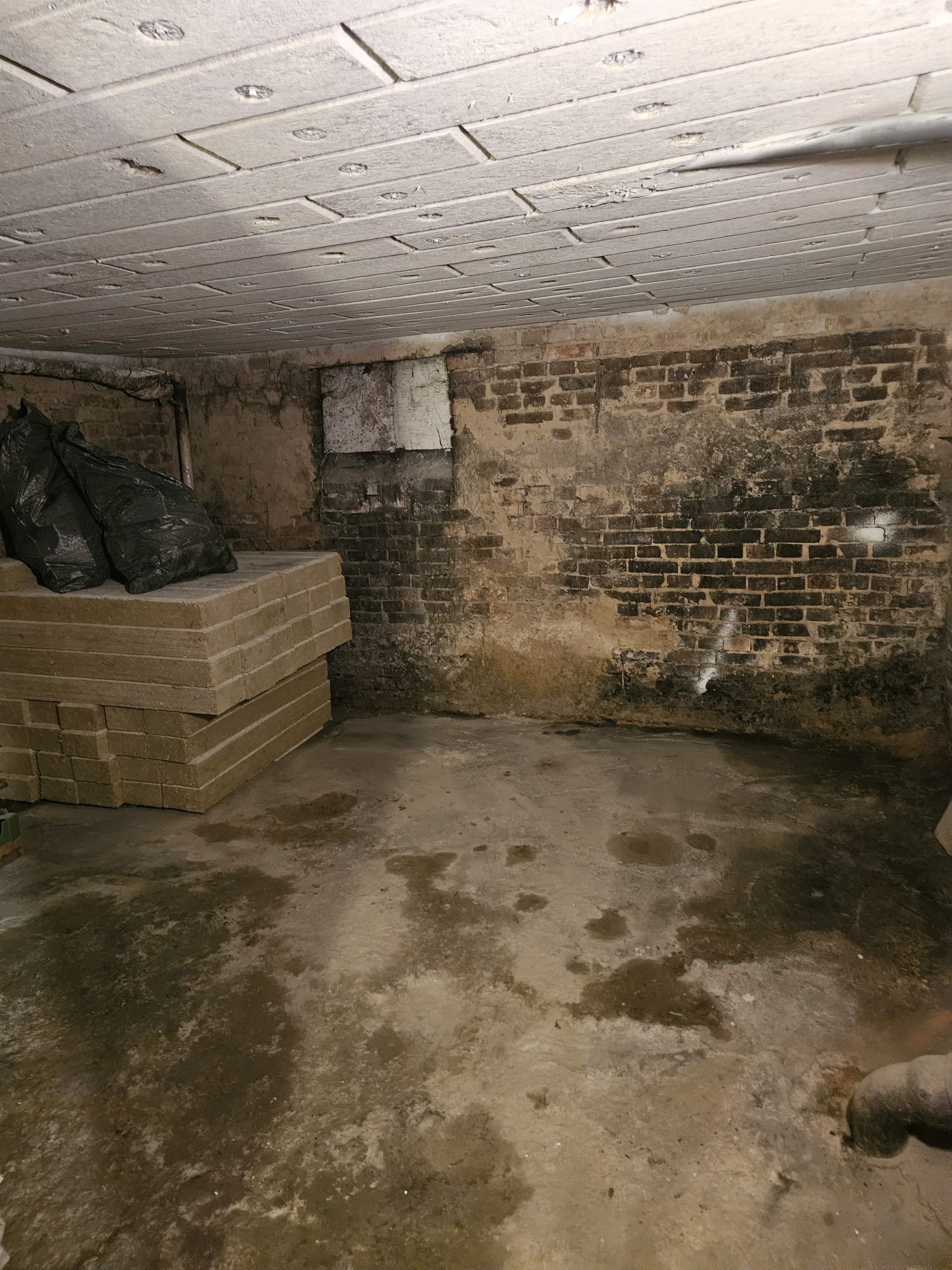 Poddasze przed ociepleniem: widoczne płyty stropowe, ceglana ściana z wilgocią, stos materiałów izolacyjnych i worki. Przygotowanie do prac izolacyjnych.