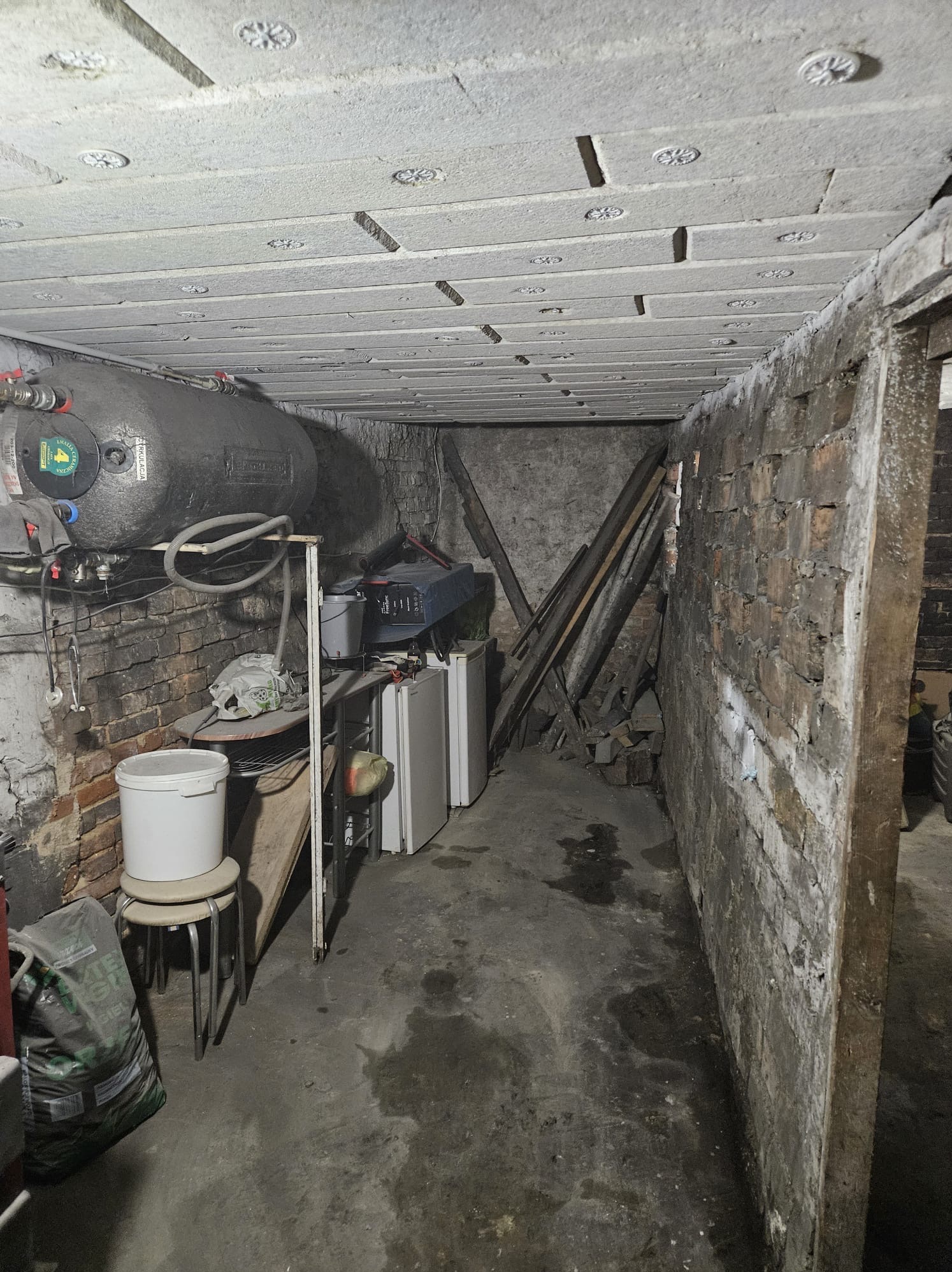 Poddasze z widoczną izolacją stropu w postaci białych paneli z dekoracyjnymi rozetami. Na ścianie zbiornik na wodę i instalacje. W tle nieporządek.