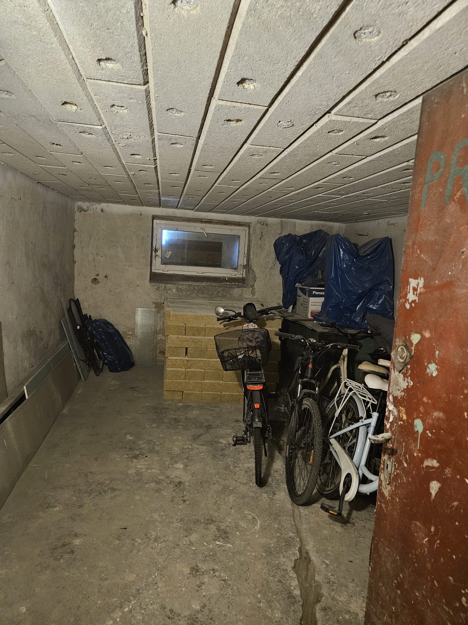 Ocieplony strop piwnicy z widocznymi płytami izolacyjnymi. W tle okno i rowery pod plandeką. Surowy stan wykończenia ścian i podłogi.