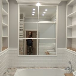 New House Project Sp. z o.o. - Wykończona łazienka z białą wanną obudowaną mozaiką, białą armaturą, lustrem z podziałami i wbudowanymi regałami; w lustrze odbija się osoba robiąca zdjęcie.