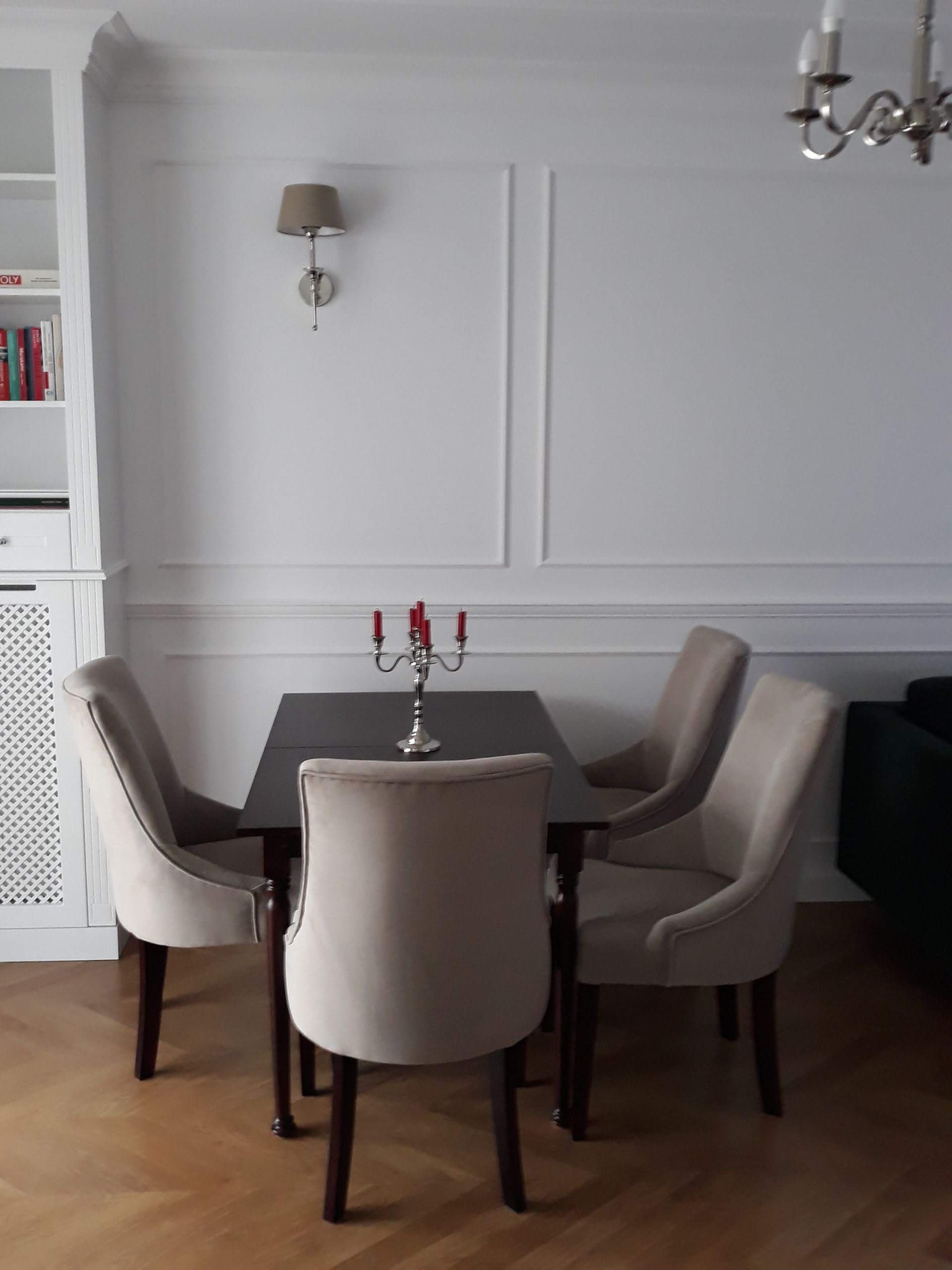 Jadalnia w jasnych barwach: ciemny stół z czterema krzesłami obitymi beżową tkaniną, srebrny świecznik z czerwonymi świecami, białe ściany z dekoracyjnymi listwami, drewniana podłoga w jodełkę...