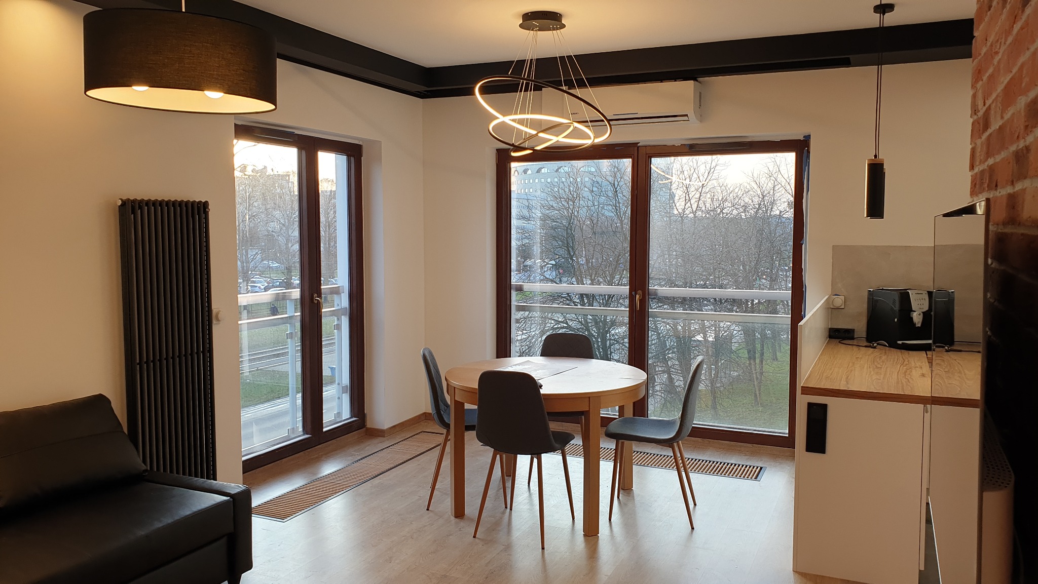 Jasne wnętrze mieszkania z okrągłym stołem, czterema krzesłami, nowoczesnym oświetleniem, widokiem z balkonu i fragmentem aneksu kuchennego z ekspressem do kawy.