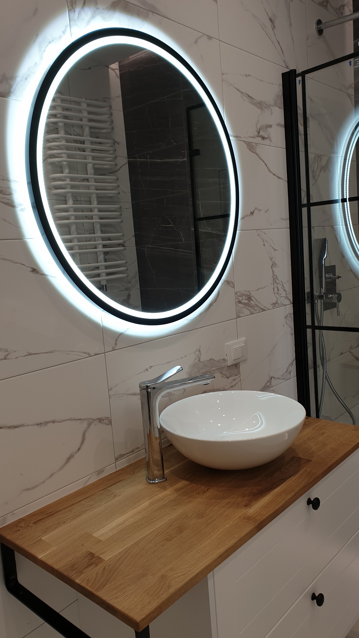 Nowoczesna łazienka z okrągłym lustrem LED, białą umywalką nablatową i drewnianym blatem szafki, marmurowe płytki na ścianie, kabina prysznicowa z czarnymi ramami w tle.
