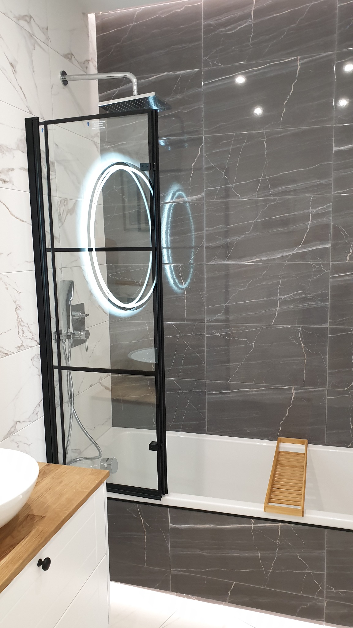 Nowoczesna łazienka z prysznicem za szklaną ścianką w czarnej ramie, marmurowe płytki w odcieniach szarości i bieli, drewniany blat pod umywalką.