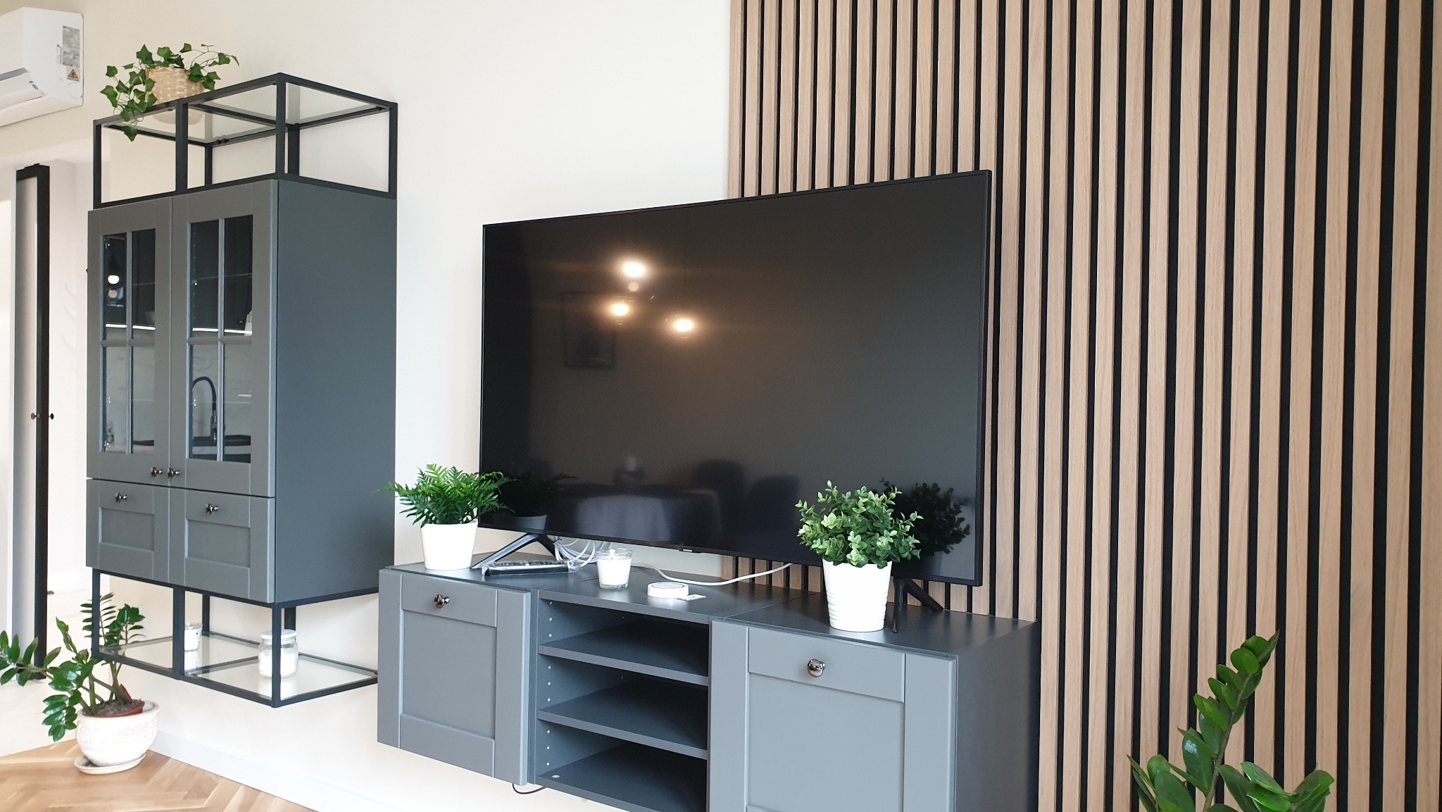 Nowoczesny salon z szarą szafką RTV, wiszącą witryną z przeszkleniami, dekoracyjną ścianą z drewnianych listew i roślinami doniczkowymi.