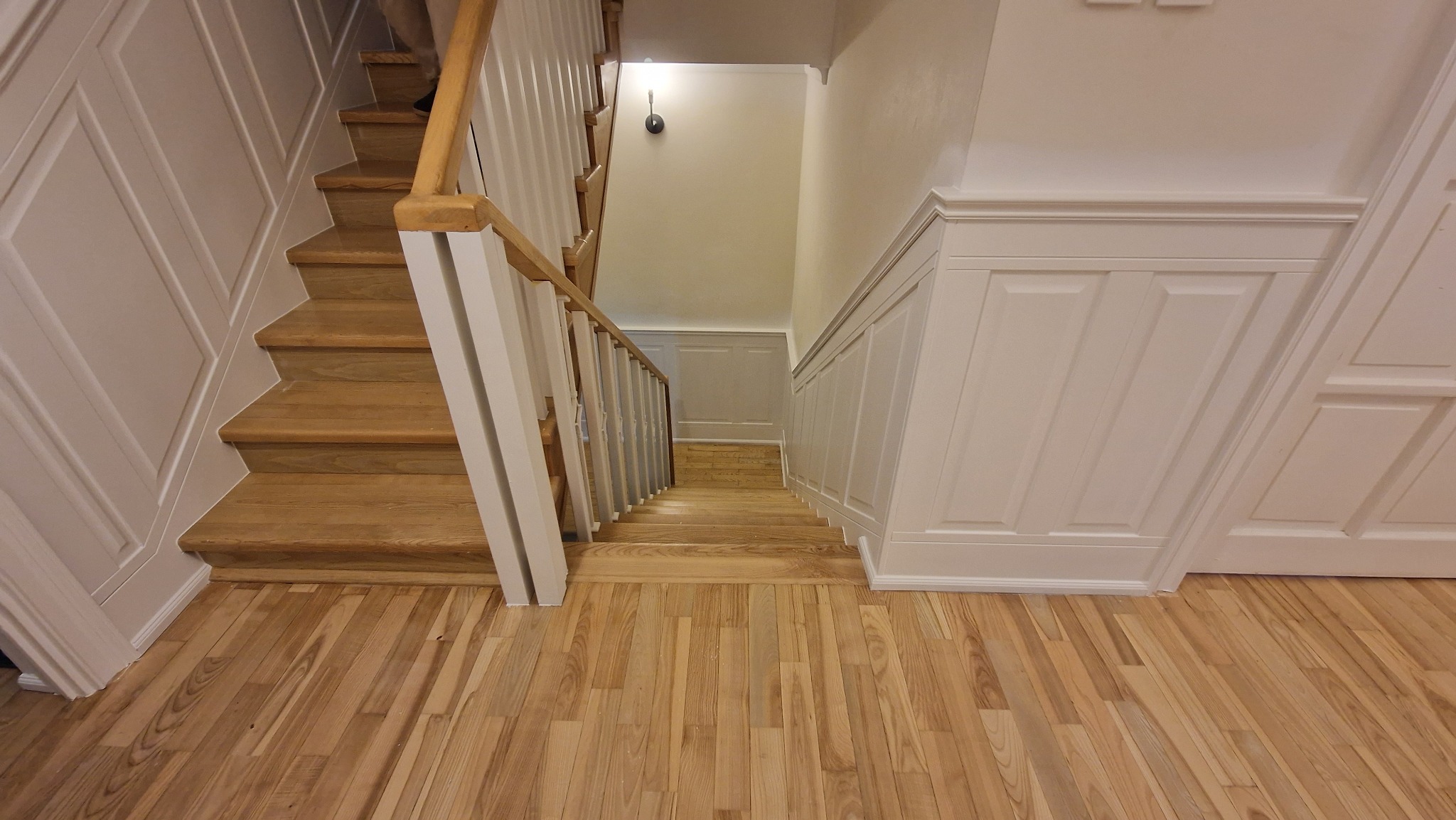 Drewniane schody z białymi balustradami i panelami ściennymi, widok z góry na dół, jasne drewno na podłodze.