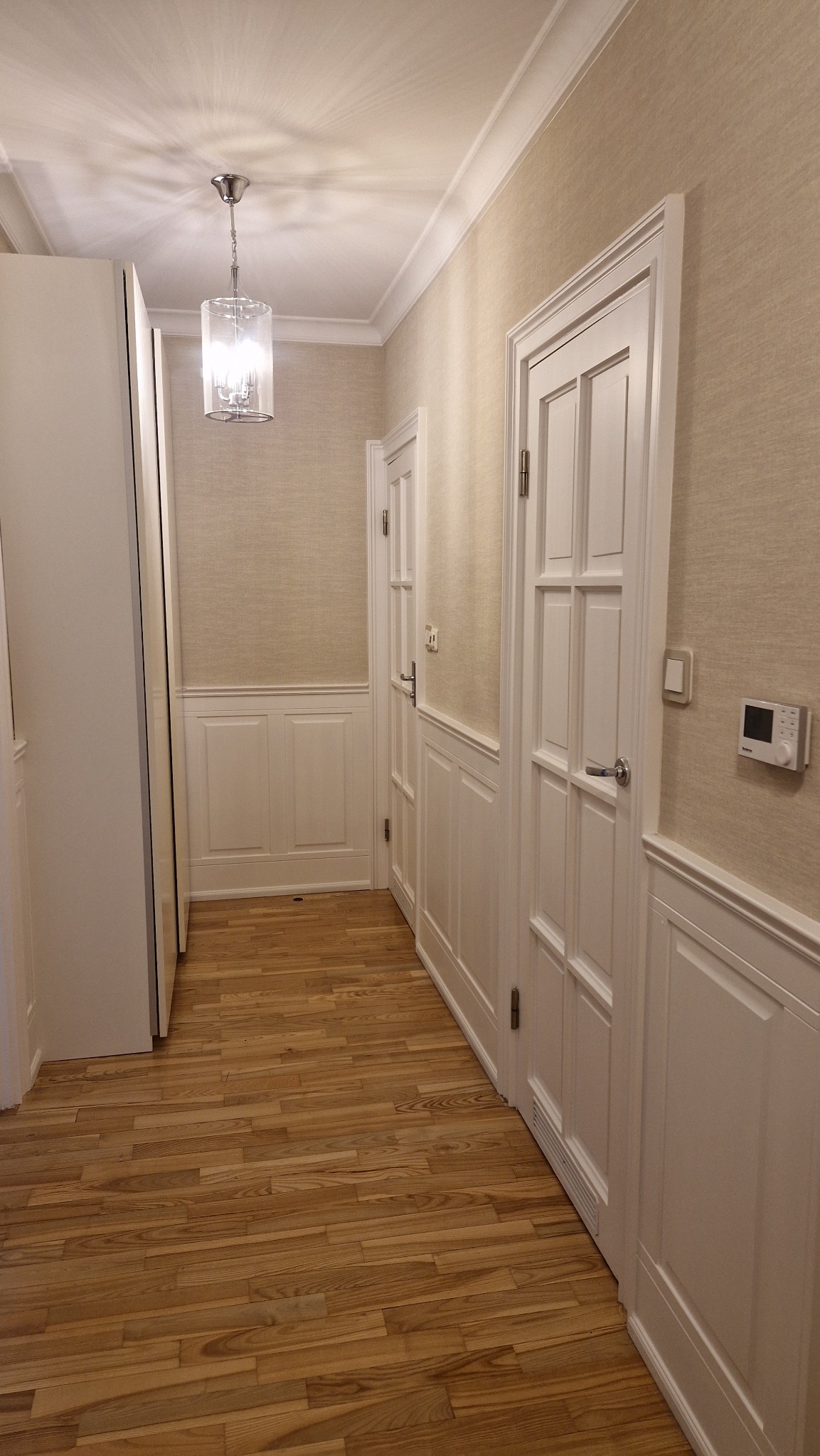Korytarz w mieszkaniu z drewnianą podłogą, białymi drzwiami panelowymi, boazerią i tapetą w neutralnym kolorze, oświetlony ozdobną lampą sufitową.