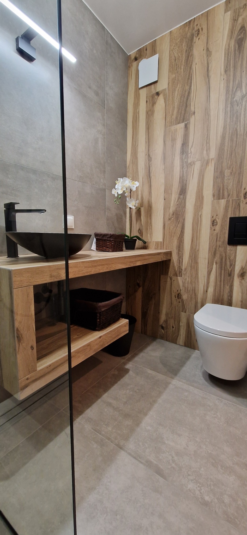 Nowoczesna łazienka z umywalką w kształcie misy na drewnianym blacie, drewnopodobnymi płytkami na ścianie i wiszącą toaletą.