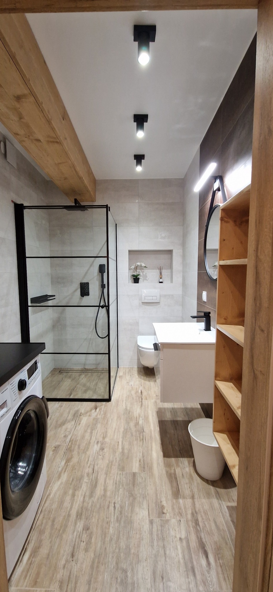 Nowoczesna łazienka z kabiną prysznicową typu walk-in z czarnymi ramami, drewnopodobnymi płytkami podłogowymi i drewnianą belką stropową, oświetleniem punktowym i białymi meblami.