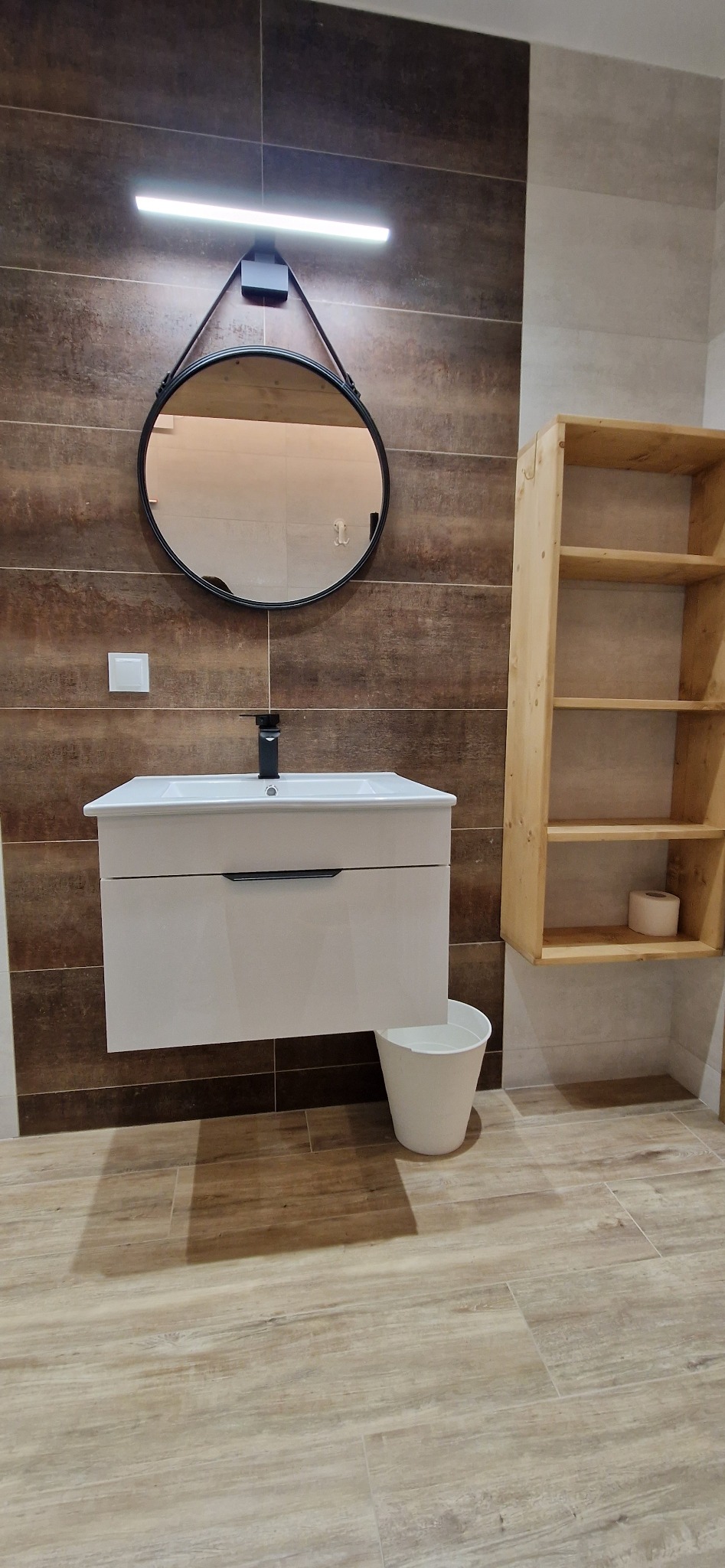 Nowoczesna łazienka z brązowymi płytkami imitującymi drewno, białą szafką pod umywalką z czarną armaturą, okrągłym lustrem z podświetleniem LED i otwartym drewnianym regałem z rolką papieru...