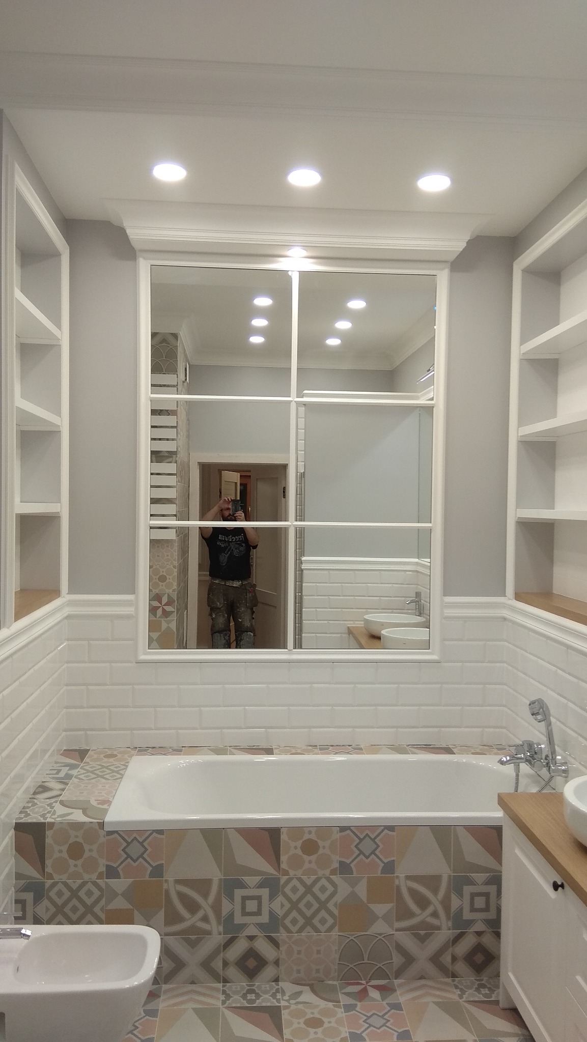 Wykończona łazienka z białą wanną obudowaną mozaiką, białą armaturą, lustrem z podziałami i wbudowanymi regałami; w lustrze odbija się osoba robiąca zdjęcie.