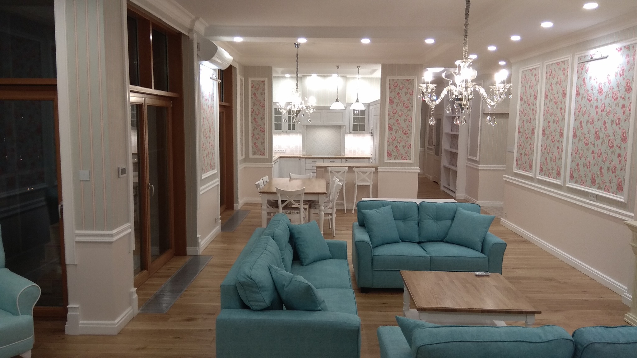 Jasne, przestronne wnętrze domu z turkusowymi sofami, drewnianą podłogą i kwiatowymi akcentami na ścianach, widok na kuchnię i jadalnię, oświetlone kryształowymi żyrandolami.