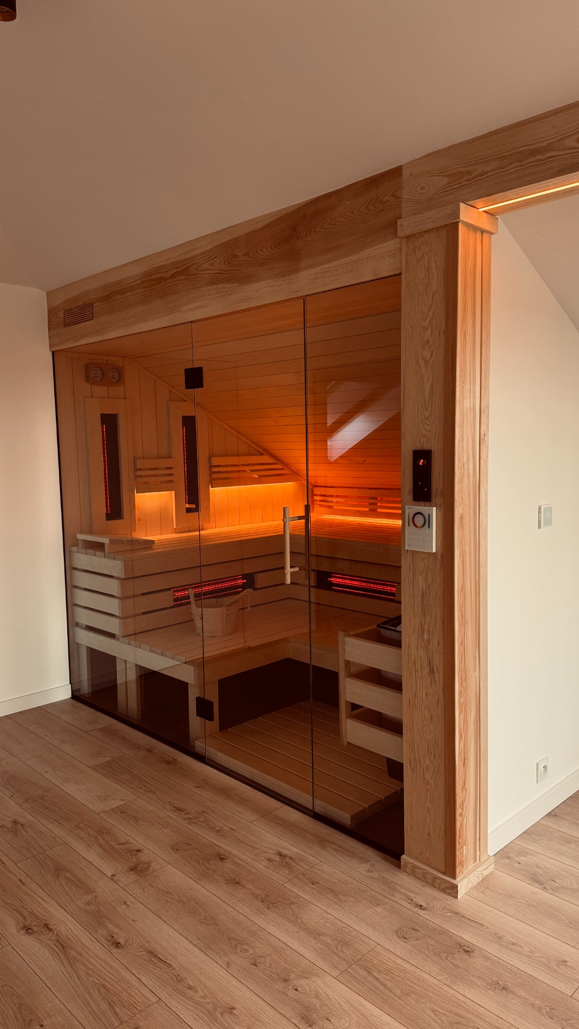 Nowoczesna sauna z przeszkloną ścianą, drewnianymi ławkami i oświetleniem LED, umieszczona w pomieszczeniu z drewnianą podłogą.