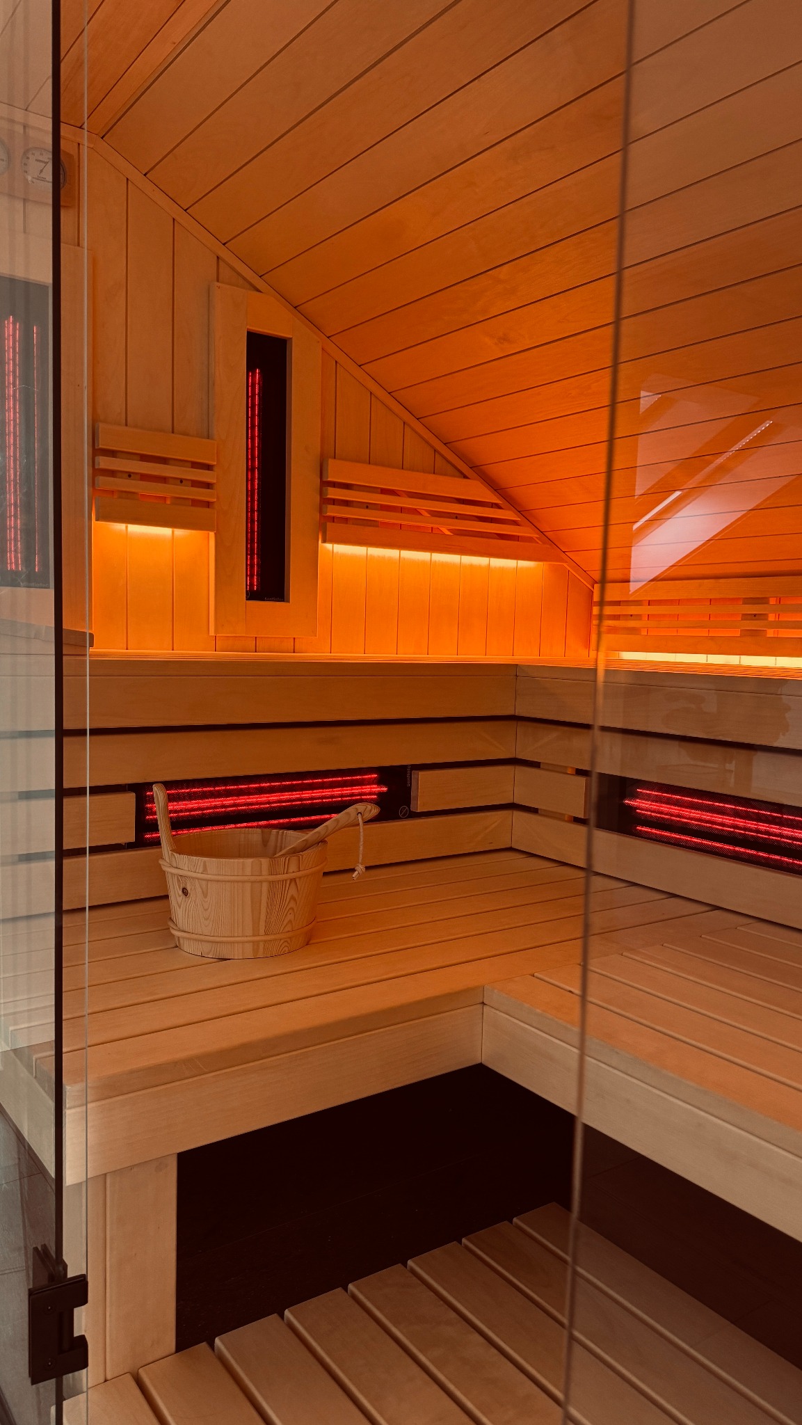 Wnętrze sauny z drewnianym wykończeniem, oświetleniem podczerwienią i wiadrem z chochlą na ławce, widok przez szklane drzwi.