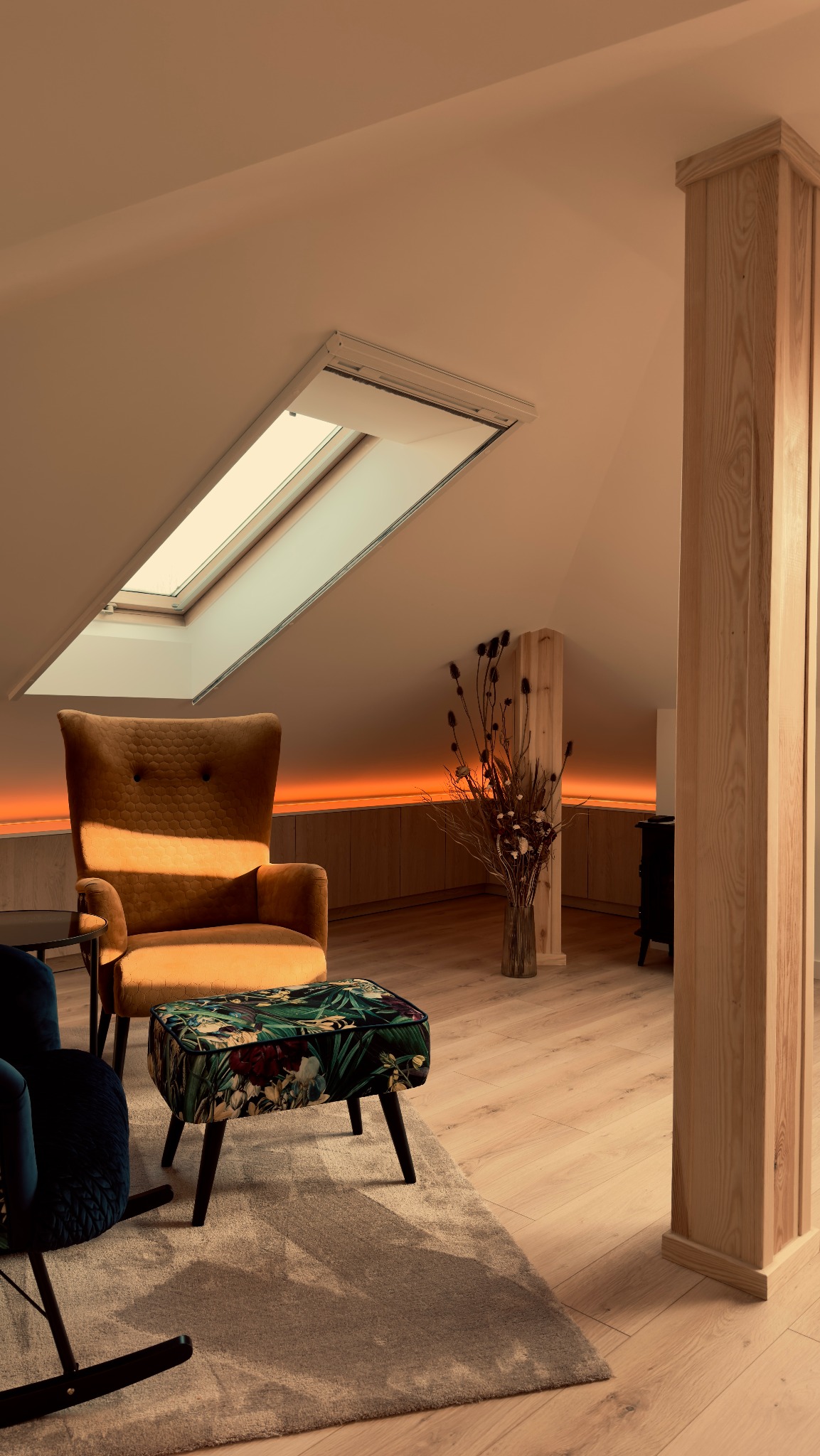 Jasne poddasze ze skośnym oknem dachowym, oświetleniem LED podszafkowym, beżowym fotelem typu uszak, pufą w tropikalne wzory i drewnianą kolumną.