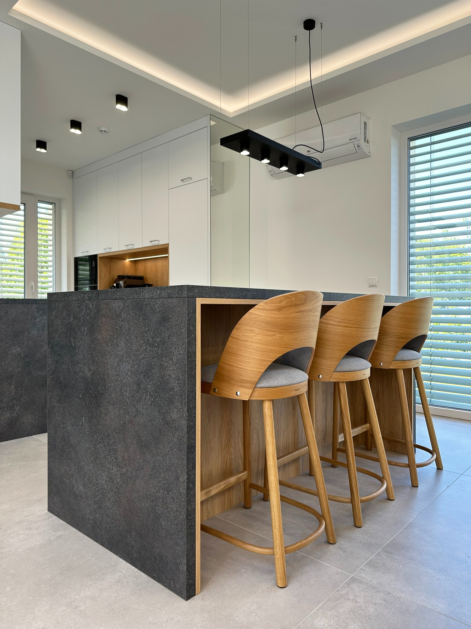 Nowoczesna kuchnia z wyspą wykończoną ciemnym kamieniem, drewniane hokery z szarymi siedziskami, białe szafki i oświetlenie punktowe.