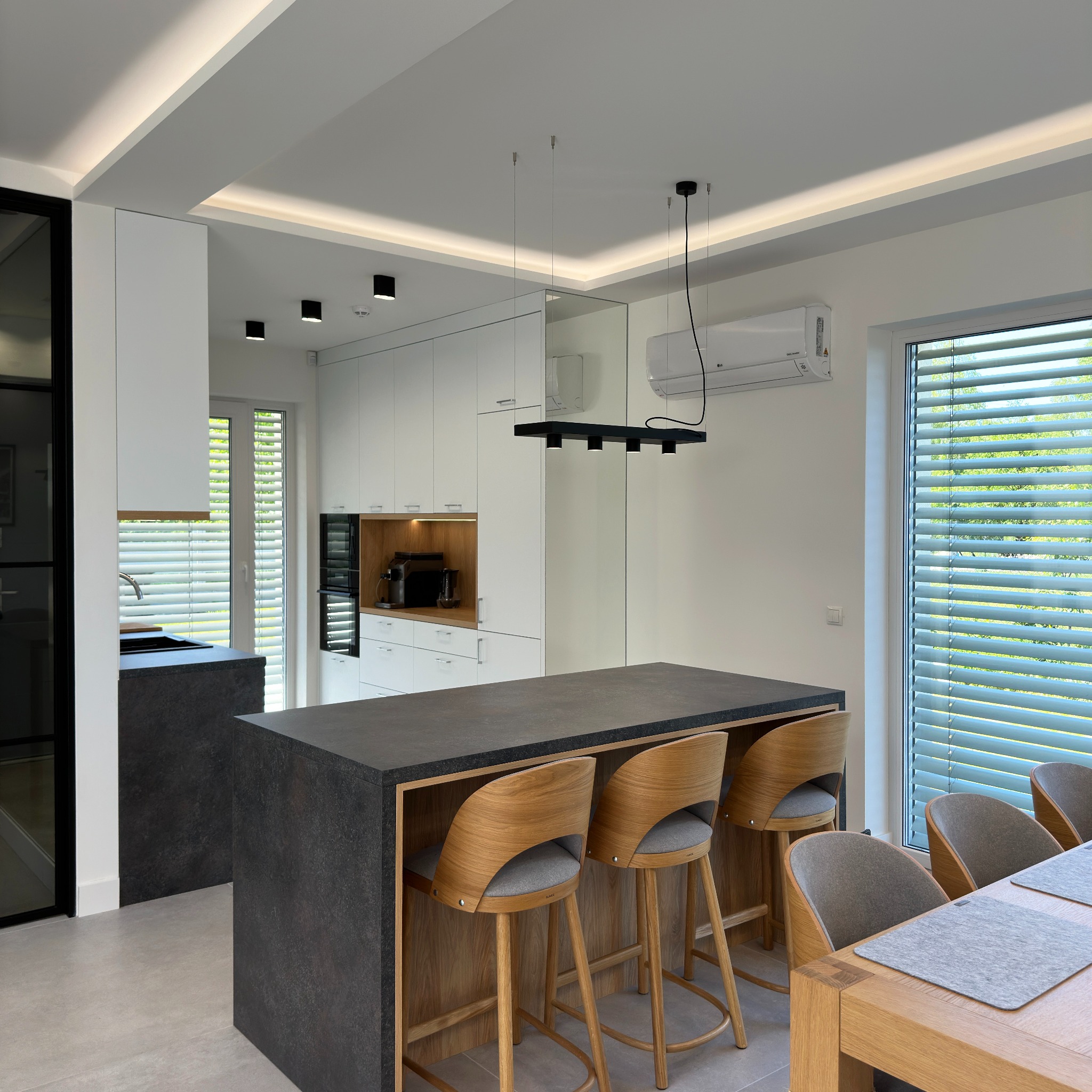 Nowoczesna kuchnia z wyspą, drewnianymi hokerami i oświetleniem LED pod sufitem, klimatyzator na ścianie.