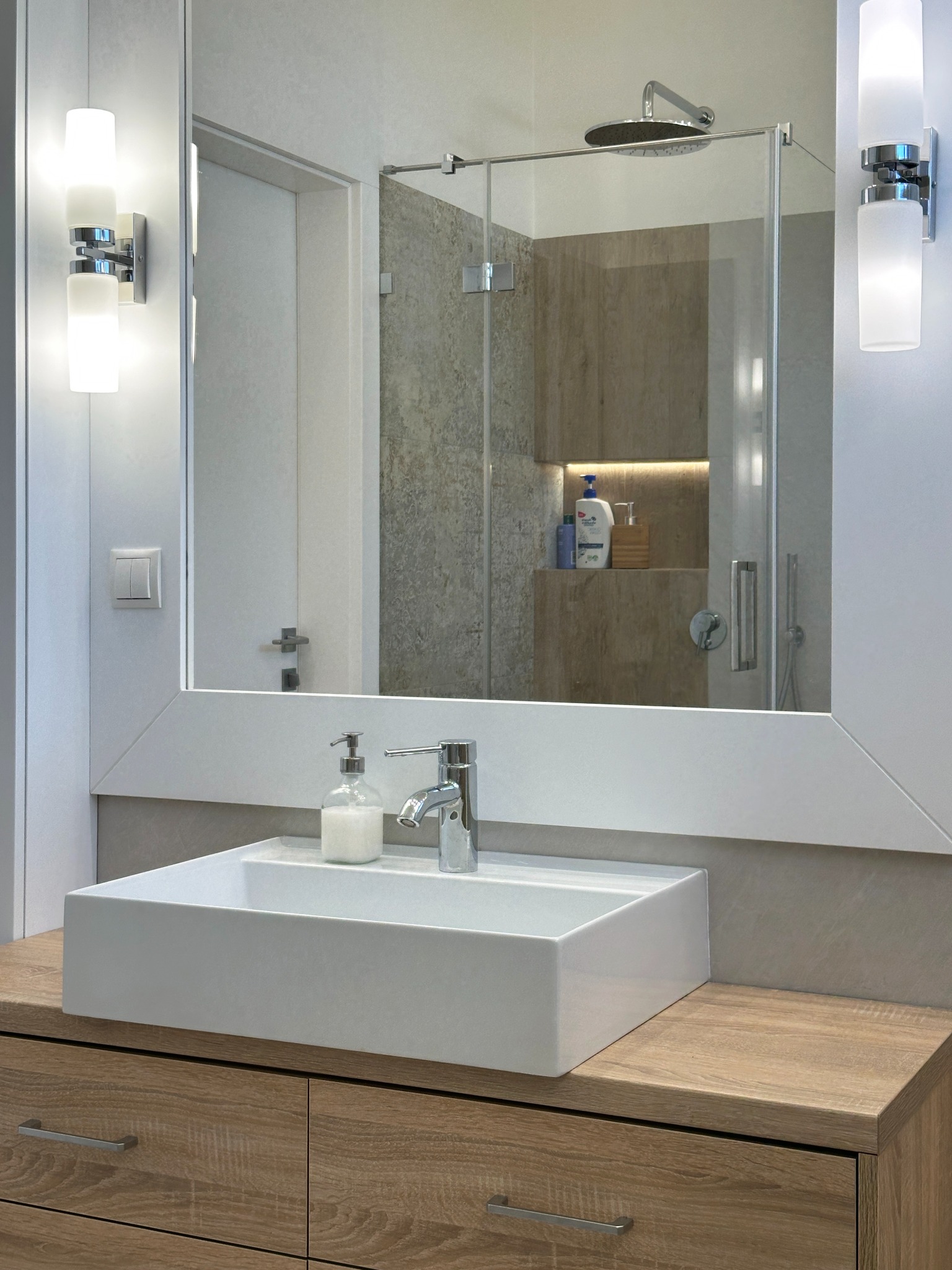 Nowoczesna łazienka z prostokątną umywalką nablatową, baterią i dozownikiem mydła, oświetlenie boczne, kabina prysznicowa z deszczownicą widoczna w lustrze.