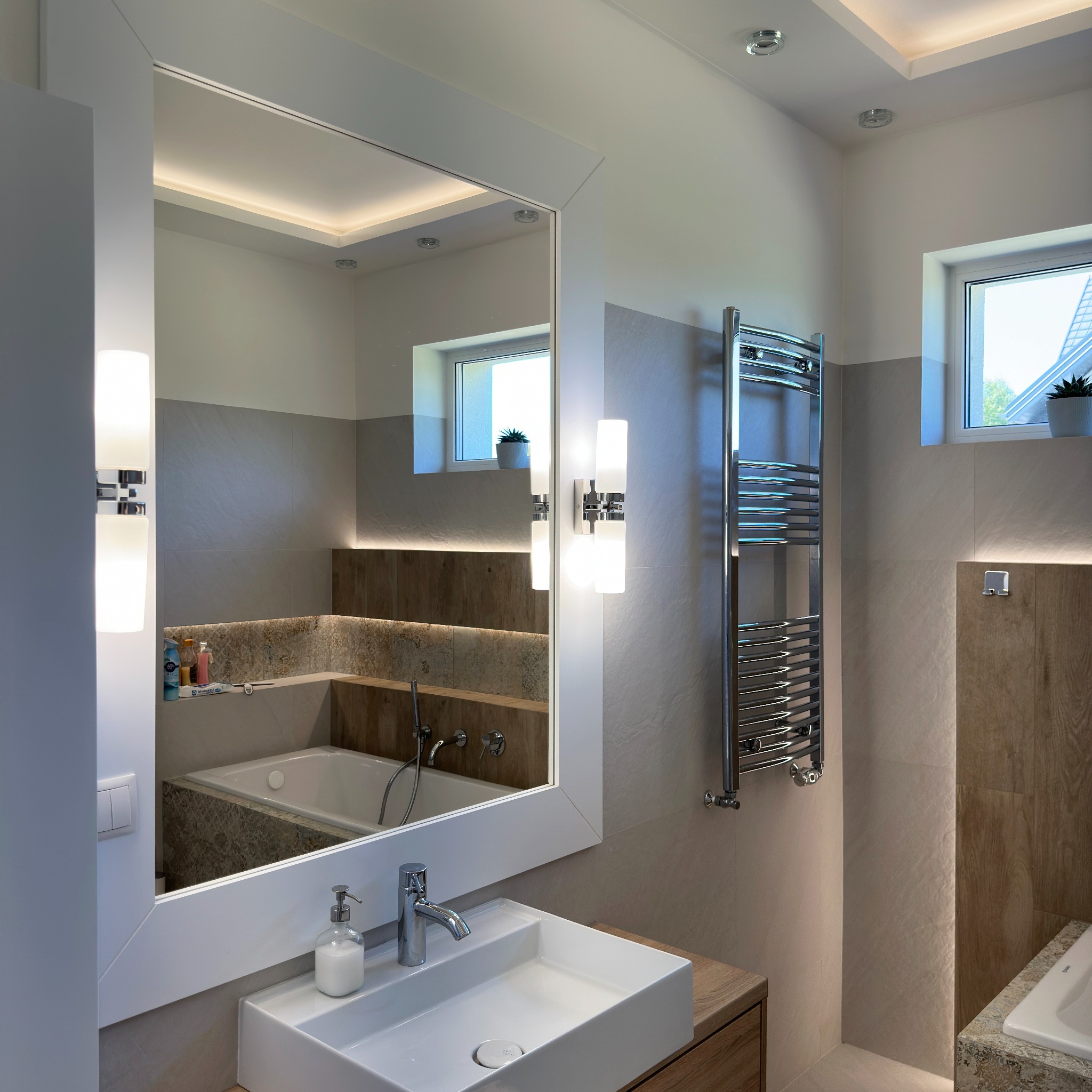 Nowoczesna łazienka z białymi płytkami, dużym lustrem z oświetleniem LED, grzejnikiem drabinkowym i umywalką nablatową, widoczna wanna w tle.