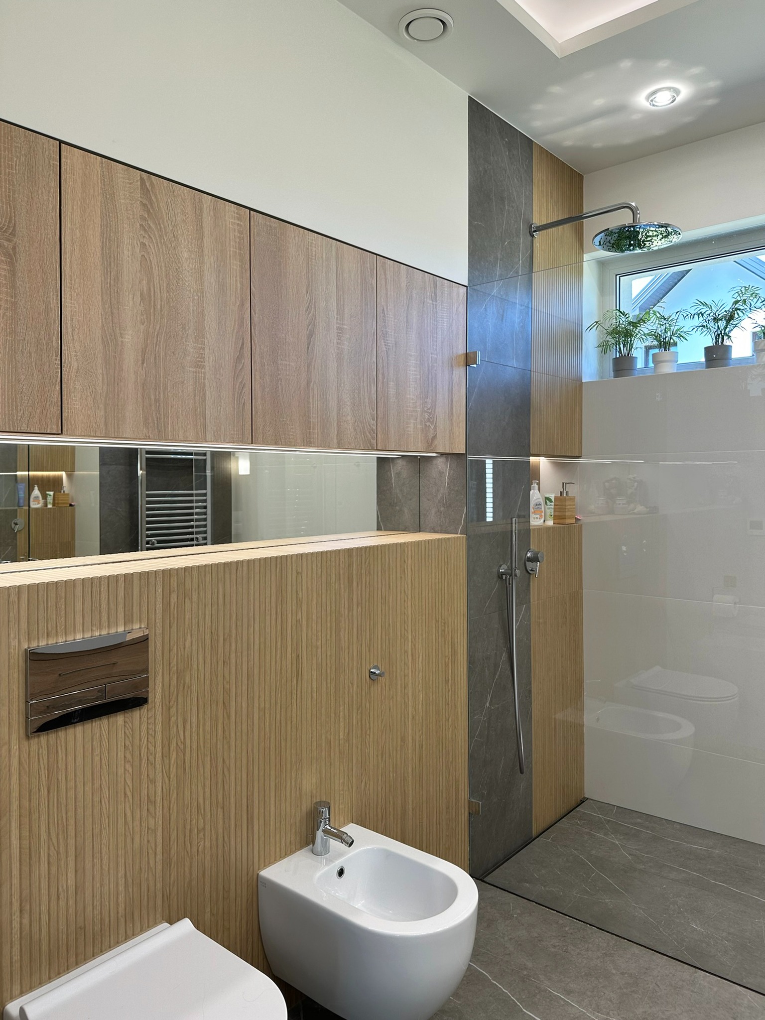 Nowoczesna łazienka z drewnianymi szafkami i panelami, szarym marmurem na podłodze i ścianie prysznica, białym bidetem i toaletą oraz zielonymi roślinami w doniczkach na parapecie okna