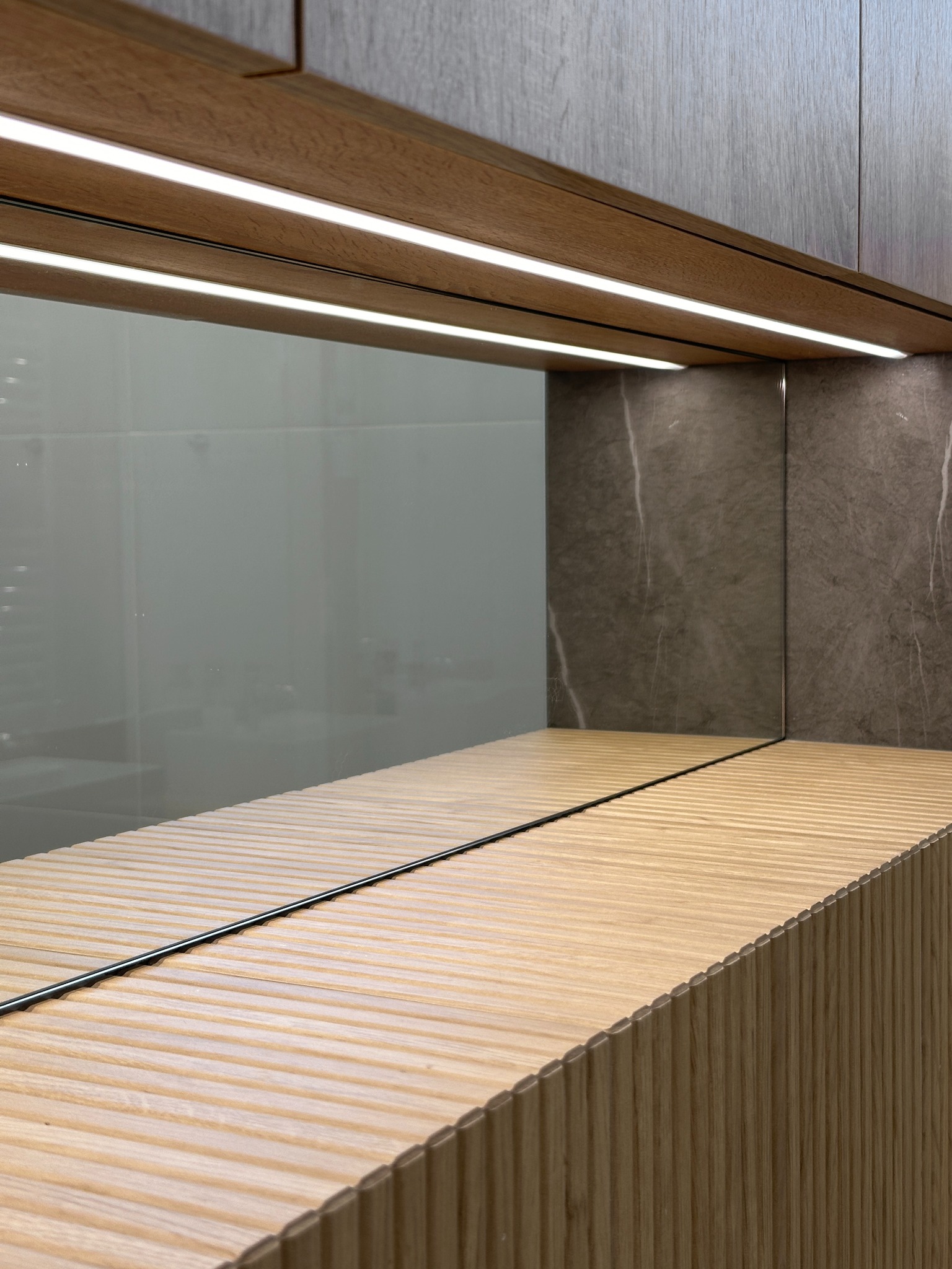 Nowoczesna łazienka: drewniana szafka umywalkowa z ryflowanym frontem, lustro z podświetleniem LED, marmurowe wykończenie ściany.