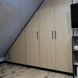 Zabudowa pod skosem z jasnego drewna z czarnymi uchwytami, dopasowana do skośnego sufitu, na podłodze parkiet z wyraźnym usłojeniem.