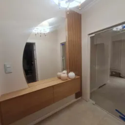 Nowoczesna aranżacja przedpokoju z owalnym lustrem, drewnianą konsolą z szufladami, dekoracyjną ścianką z drewnianych listewek i szklanymi drzwiami do garderoby.