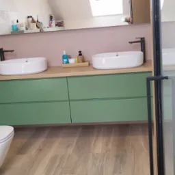 Nowoczesna łazienka z zielonymi szafkami pod umywalkami, drewnianym blatem, białymi umywalkami nablatowymi i wiszącą toaletą.