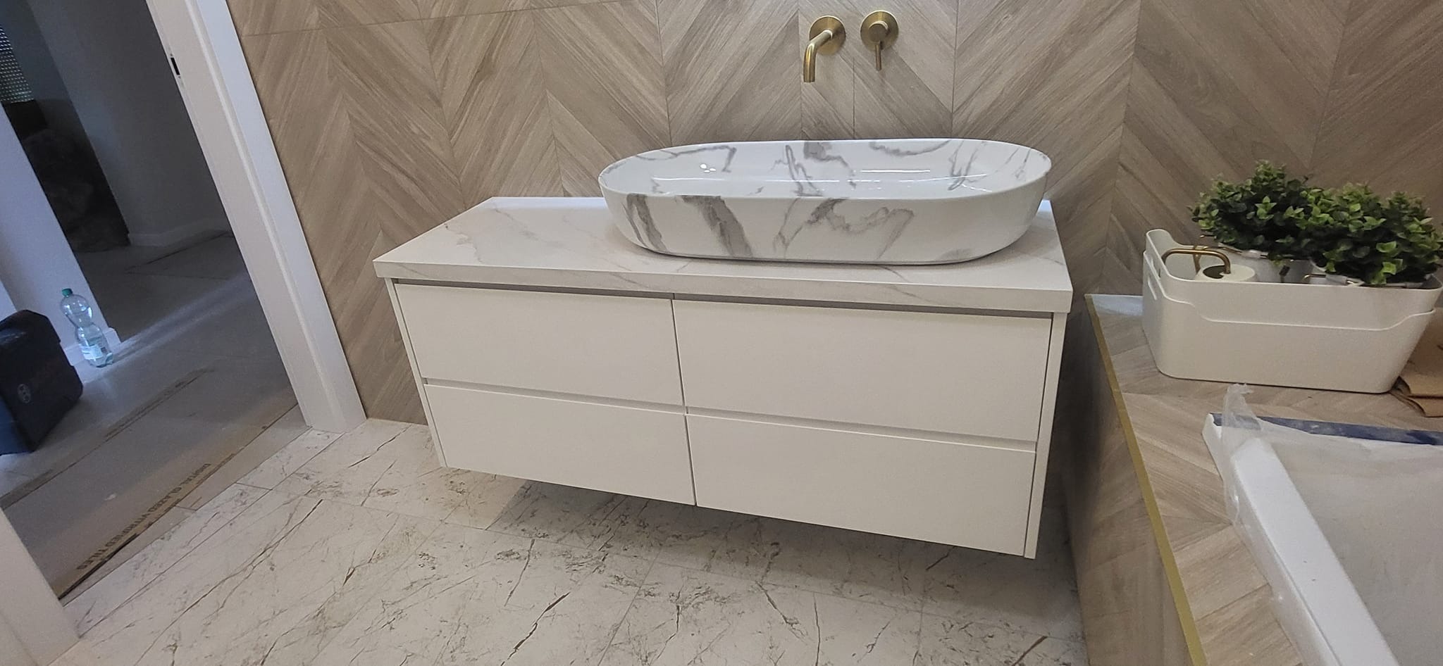 Nowoczesna łazienka z białą szafką pod umywalkę z marmurowym blatem i umywalką nablatową, złota armatura, płytki imitujące drewno ułożone w jodełkę na ścianie i podłodze.