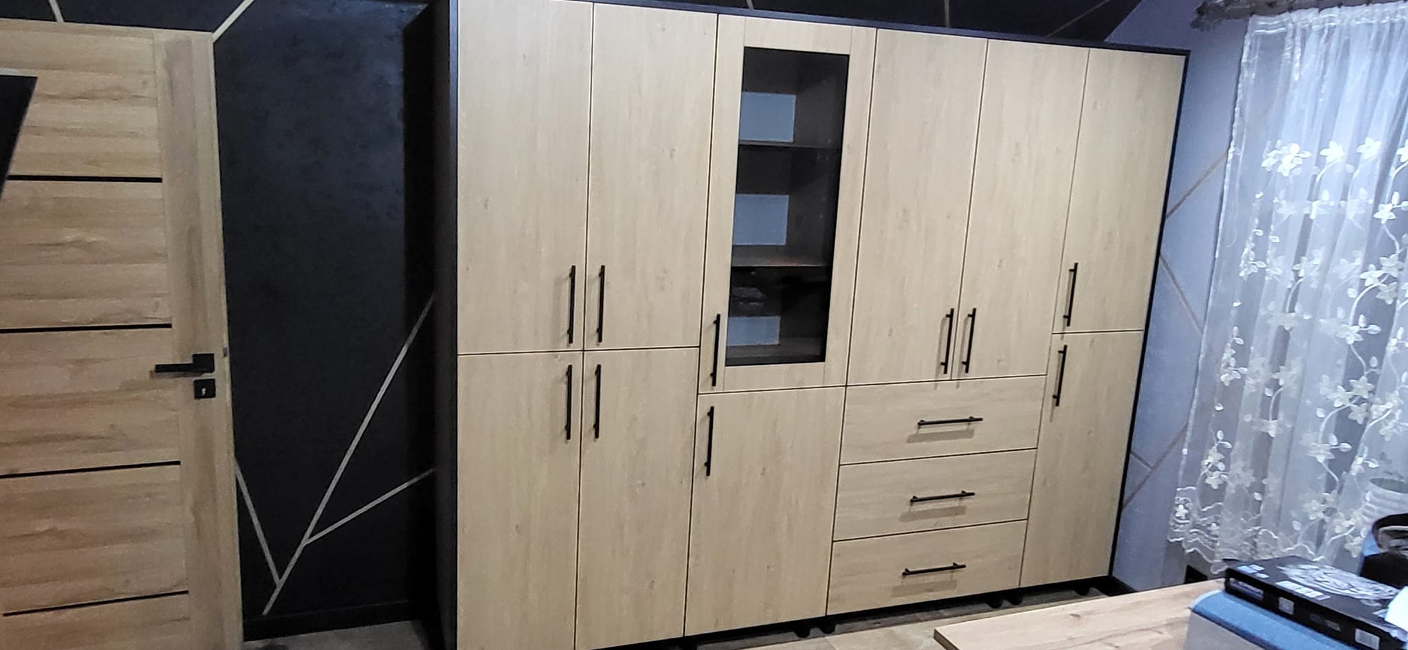 Jasna zabudowa meblowa z ciemnymi uchwytami, z szafką z przeszklonym frontem i szufladami, na tle czarnej ściany z geometrycznym wzorem i zasłony z motywem kwiatowym.