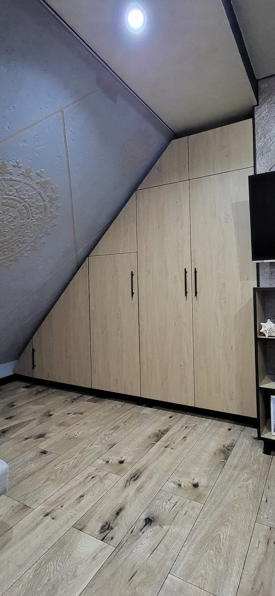 Zabudowa pod skosem z jasnego drewna z czarnymi uchwytami, dopasowana do skośnego sufitu, na podłodze parkiet z wyraźnym usłojeniem.