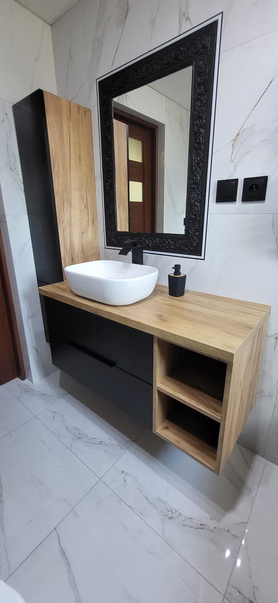 Nowoczesna łazienka z wiszącą szafką pod umywalkę z drewnianym blatem i czarnymi frontami, owalna biała umywalka nablatowa, czarna bateria i ozdobne lustro w czarnej ramie.