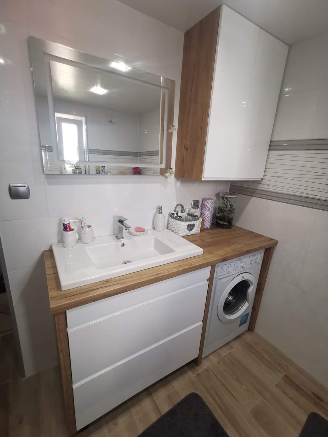 Nowoczesna łazienka z białą umywalką wpuszczoną w drewniany blat, szafką z szufladami i pralką w zabudowie, wisząca szafka z białymi frontami i drewnianym bokiem, lustro z podświetleniem LED.
